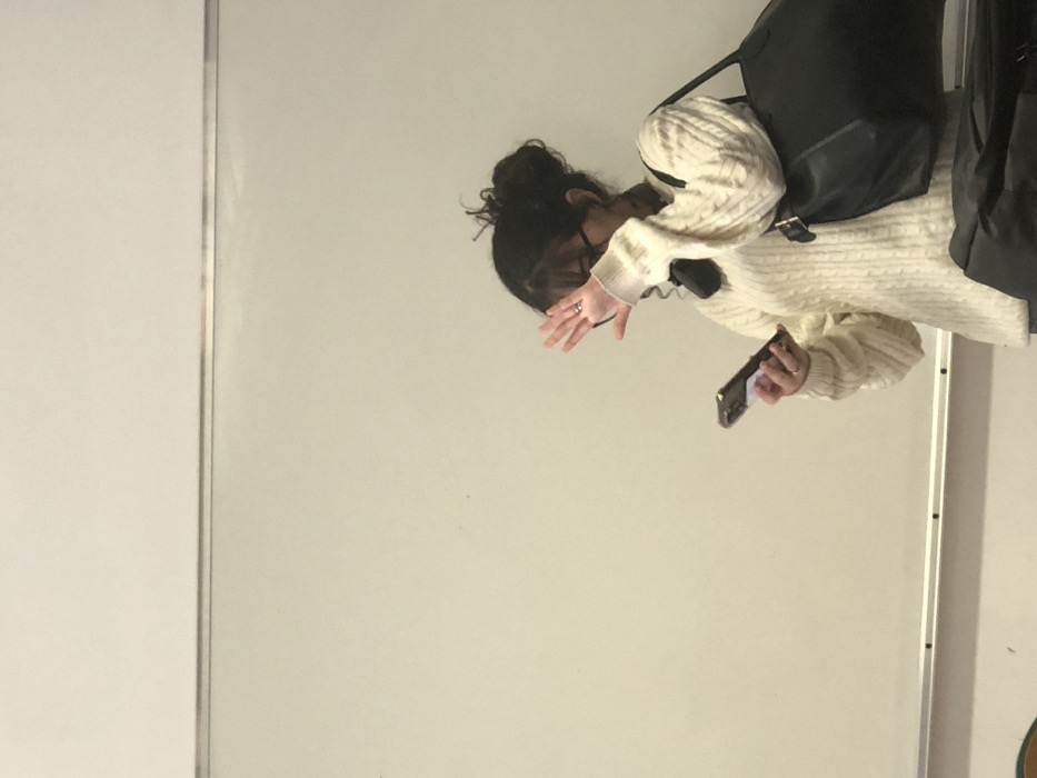 The scene is an interior location in Schiltigheim, France, characterized by a large white board against a light-colored wall, consistent with a classroom or meeting room. A female individual is captured from the mid-torso upward, positioned facing the viewer. Her dark, curly hair is gathered into a messy bun, and she wears eyeglasses. She is dressed in a cream-colored cable-knit sweater, with a dark accessory, possibly headphones, visible around her neck. A metallic ring adorns the fourth finger of her left hand. She holds a dark-cased smartphone with a white attachment on its reverse side in her left hand, directed downwards. Her right hand is raised, palm open, positioned in front of her face, partially obscuring her facial features. A large black shoulder bag is carried on her right side. Partial views of a brown, possibly wooden, chair are visible in the lower left, and a dark bag or object is present in the lower right portion of the frame.