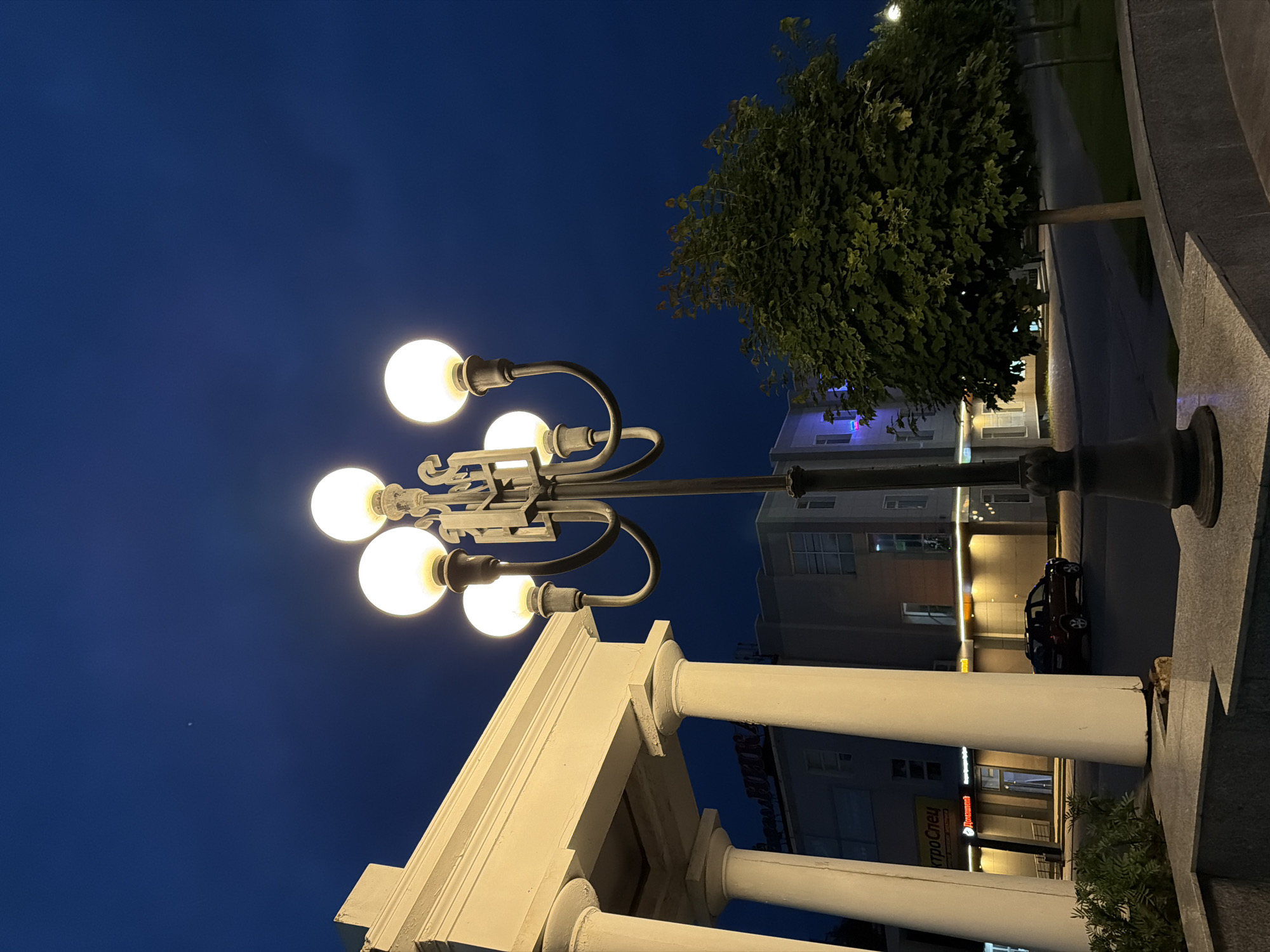 The image captures a street scene in Chaikovsky, Russia, during twilight. The foreground features a light post with multiple illuminated globes, casting a warm glow against the darkening sky. To the left, a portion of a building with white columns and an overhang is visible. In the mid-ground, a dark-colored SUV is parked on the street in front of a building with illuminated windows and signage. Further back, a lush green tree stands prominently on the right side of the frame, with other trees lining the street. The sky is a deep blue, indicating the transition from day to night, and a faint star or distant light is visible high in the sky.