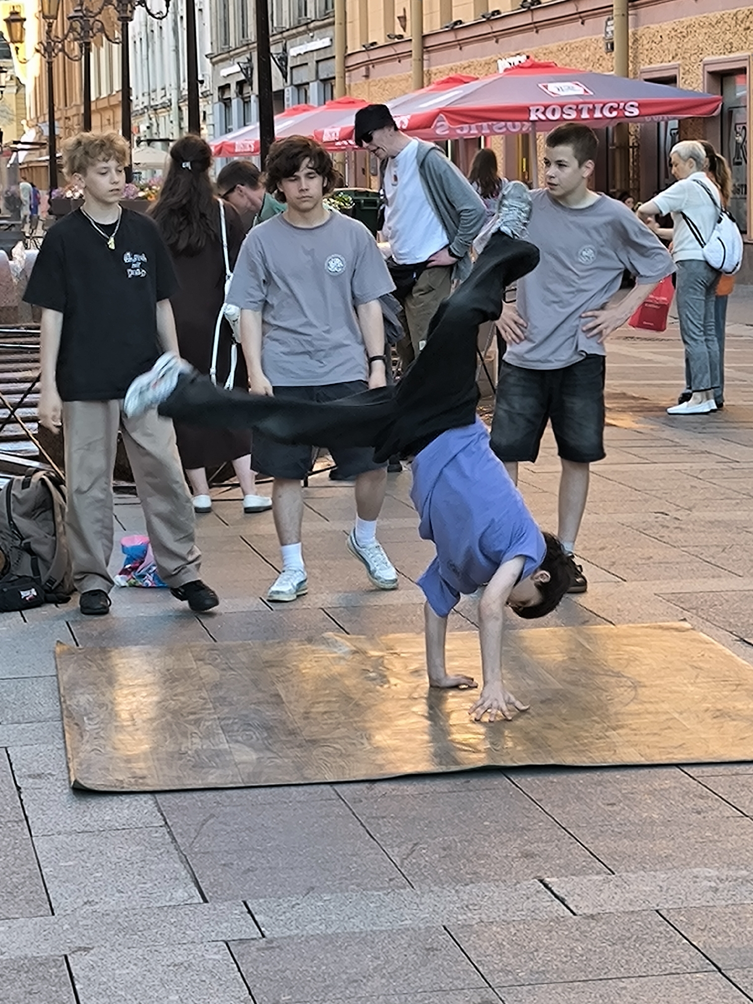 The image depicts a street scene in Saint Petersburg, Russia, featuring a young individual performing a one-handed handstand on a rectangular, reflective mat placed on a paved pedestrian thoroughfare. The performer, clad in a blue short-sleeved shirt and dark pants, is balanced on their left hand, with legs extended upwards and head facing the ground.
Surrounding the performer are several bystanders. To the left, a young male with curly blonde hair wears a black t-shirt and light brown pants, standing with his hands at his sides. A dark backpack rests on the pavement near his left foot. To the performer's right, a young male with dark curly hair, wearing a gray t-shirt and dark shorts, stands with his hands on his hips, observing the performance. Further back on the right, an older female with light-colored hair, dressed in a white shirt and gray pants, carries a white shoulder bag and looks towards the left. A red shopping bag is near her feet. Other indistinct figures are visible in the background.
The ground is paved with light gray rectangular stone slabs. In the background, multi-story buildings with classic architectural details line the street. A prominent red and gray umbrella bearing the white "ROSTIC'S" logo is visible on the right, indicating a fast-food establishment. Ornate black lampposts are present on the left side of the street. The scene is well-lit, suggesting daylight conditions.