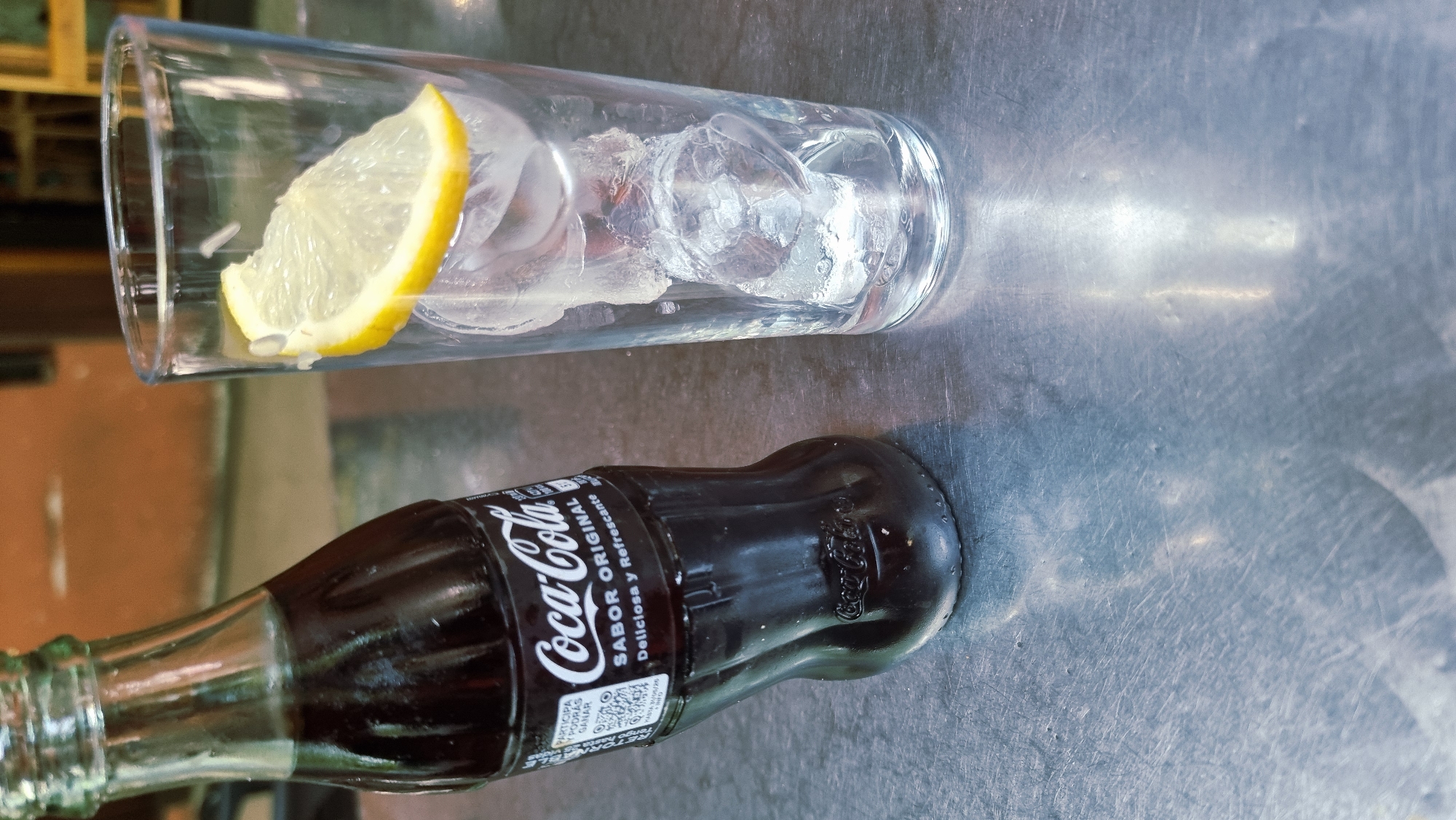 A glass with ice and a slice of lemon sits on a metal surface. Next to it, a dark brown glass bottle of Coca-Cola is lying on its side. The bottle has a label with white text that reads "Coca-Cola" and "SABOR ORIGINAL." The metal surface appears to be a table or countertop, with reflections and scuffs visible. The lighting suggests it might be daytime, possibly indoors or in a shaded outdoor area. The overall scene conveys a sense of refreshment and casual enjoyment, with the Coca-Cola bottle implying a drink is being served or consumed. The location context is identified as Olot, Spain.