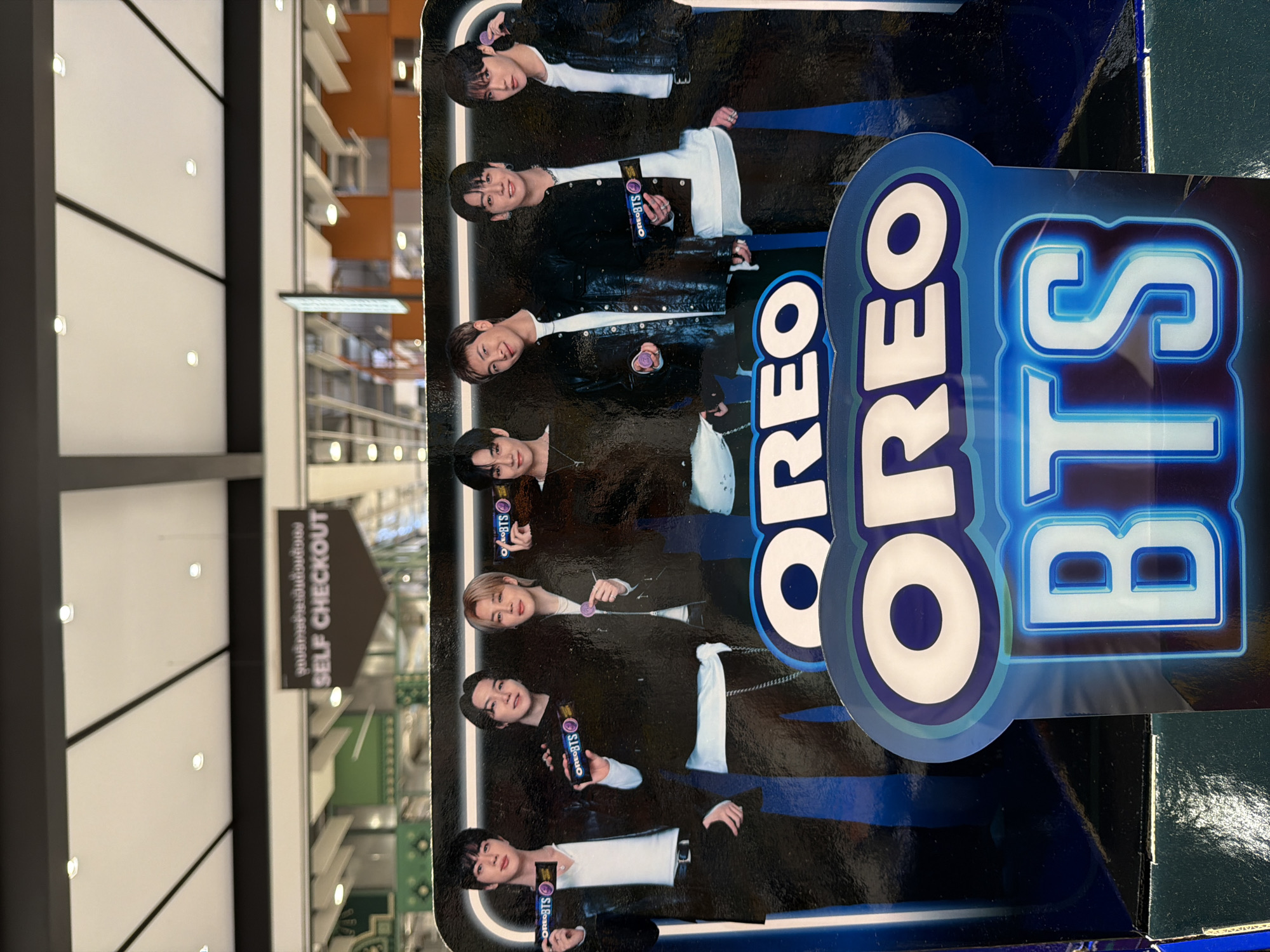 The image shows a promotional display for Oreo BTS cookies, featuring life-sized cardboard cutouts of seven men, presumably members of the K-pop group BTS. They are dressed in dark clothing and holding Oreo products. The display is positioned in front of a blurred background that appears to be an indoor retail environment with ceiling lights visible. A sign with Thai text and "SELF CHECKOUT" in English is visible in the upper portion of the image, suggesting the location is a store or supermarket. The overall impression is a vibrant commercial setting.