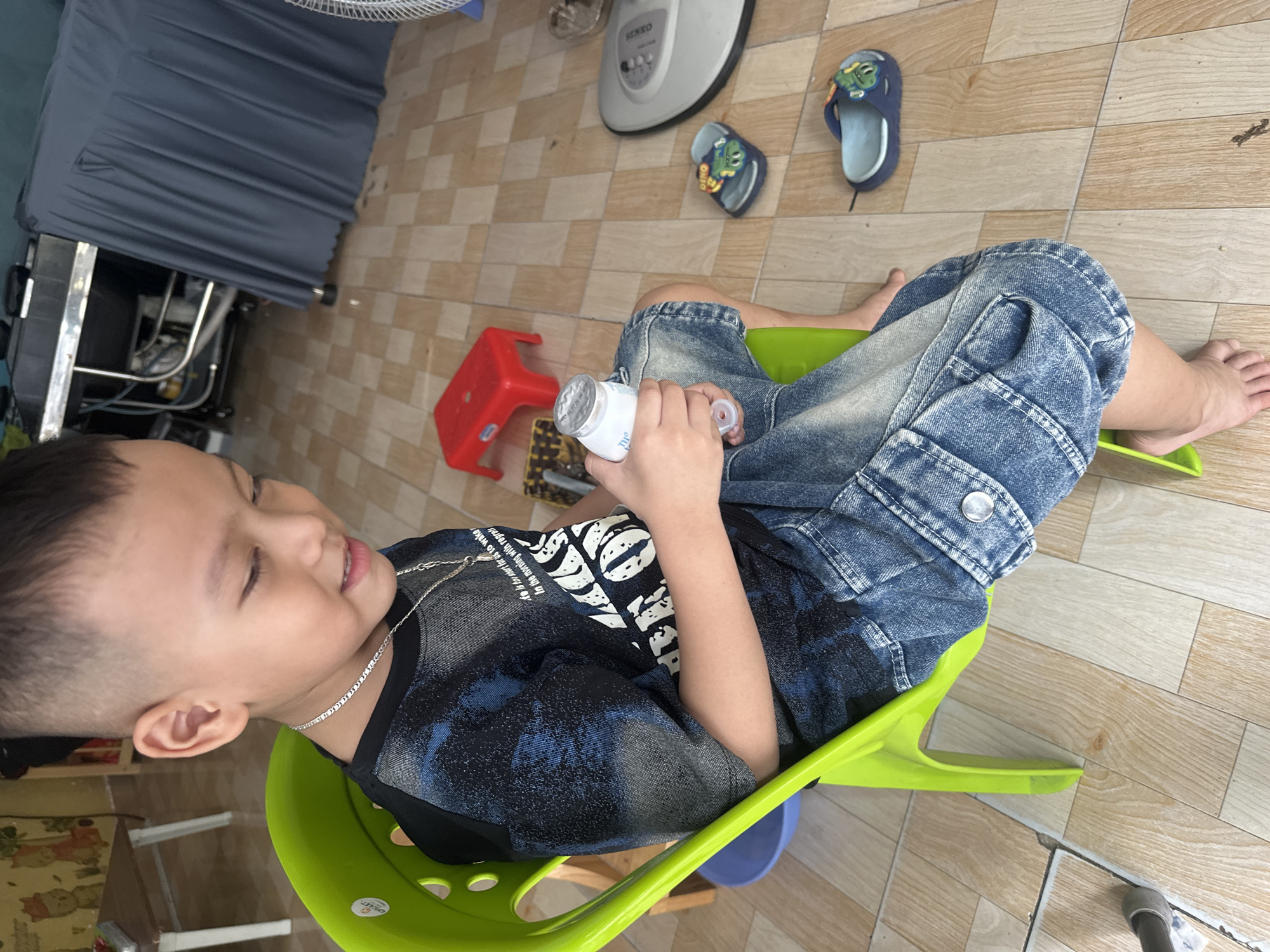 A boy is sitting in a green chair, holding a white bottle, located in the city of Bac Nha Trang Ward, Vietnam. The boy has a short haircut and wears a silver necklace. He is wearing a blue and black patterned shirt and denim shorts. A small red stool is on the floor next to him. In the background, there are children's sandals and a white fan. The floor is tiled.