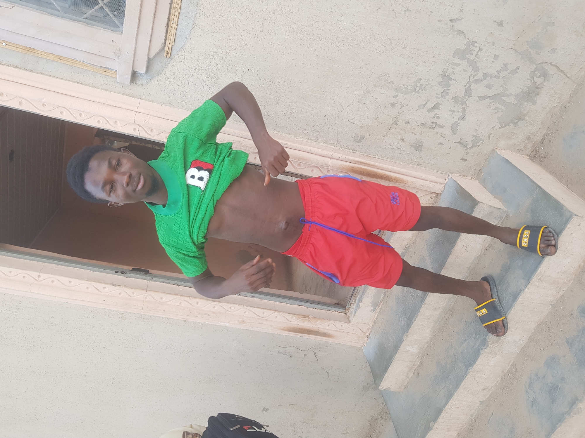 A young man stands on concrete steps in Dutsinma B, Nigeria. He is wearing a green shirt, red shorts, and Zara brand sandals. His shirt is lifted, exposing his abdomen. He faces the camera. The background features a door frame, window, and textured wall. A partial view of a black bag is visible at the bottom of the frame. The scene is well-lit.