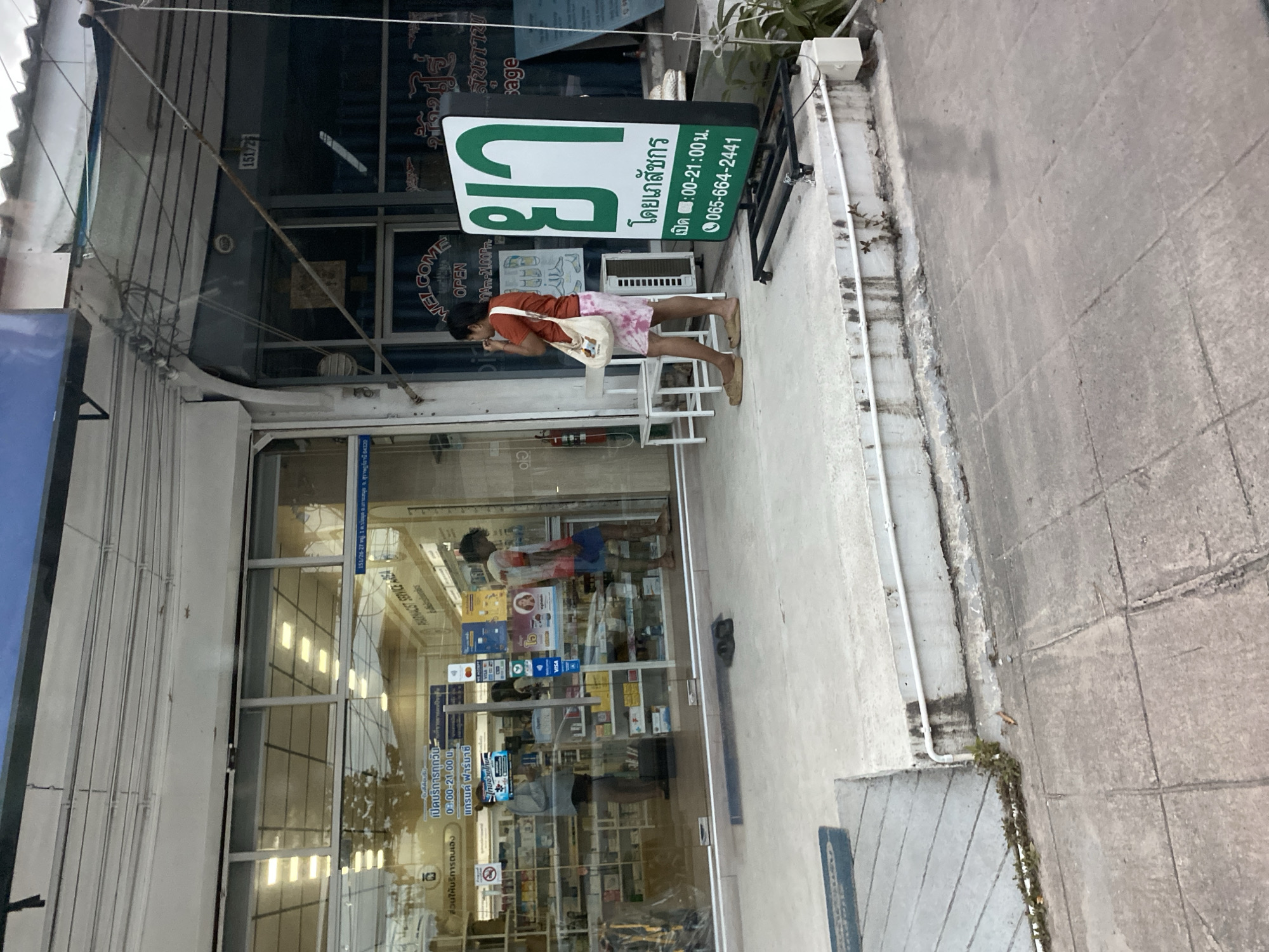 A person wearing an orange shirt and patterned shorts stands on a sidewalk in front of a pharmacy. They are holding a white tote bag over their shoulder and appear to be looking down at their hands. The pharmacy has a large glass storefront with displays of products visible inside. A green and white sign above the entrance reads "ยา" (Ya) which means medicine in Thai, and also "โดยเภสัชกร" (Doi Phenachakorn) meaning "By Pharmacist". Below this, it lists operating hours as "09:00-21:00 น." and a phone number. Another sign on the storefront says "WELCOME OPEN 09.00-21.00 น.". The scene is set in an outdoor environment with a concrete sidewalk and a building facade. The lighting suggests it might be daytime. The overall atmosphere appears to be casual and everyday. The location is identified as Baan Bo Phut, Thailand, with text on a sign confirming "ต.บ่อผุด อ.เกาะสมุย จ. สุราษฎร์รานี 84320" which translates to Bo Phut Subdistrict, Koh Samui District, Surat Thani Province.