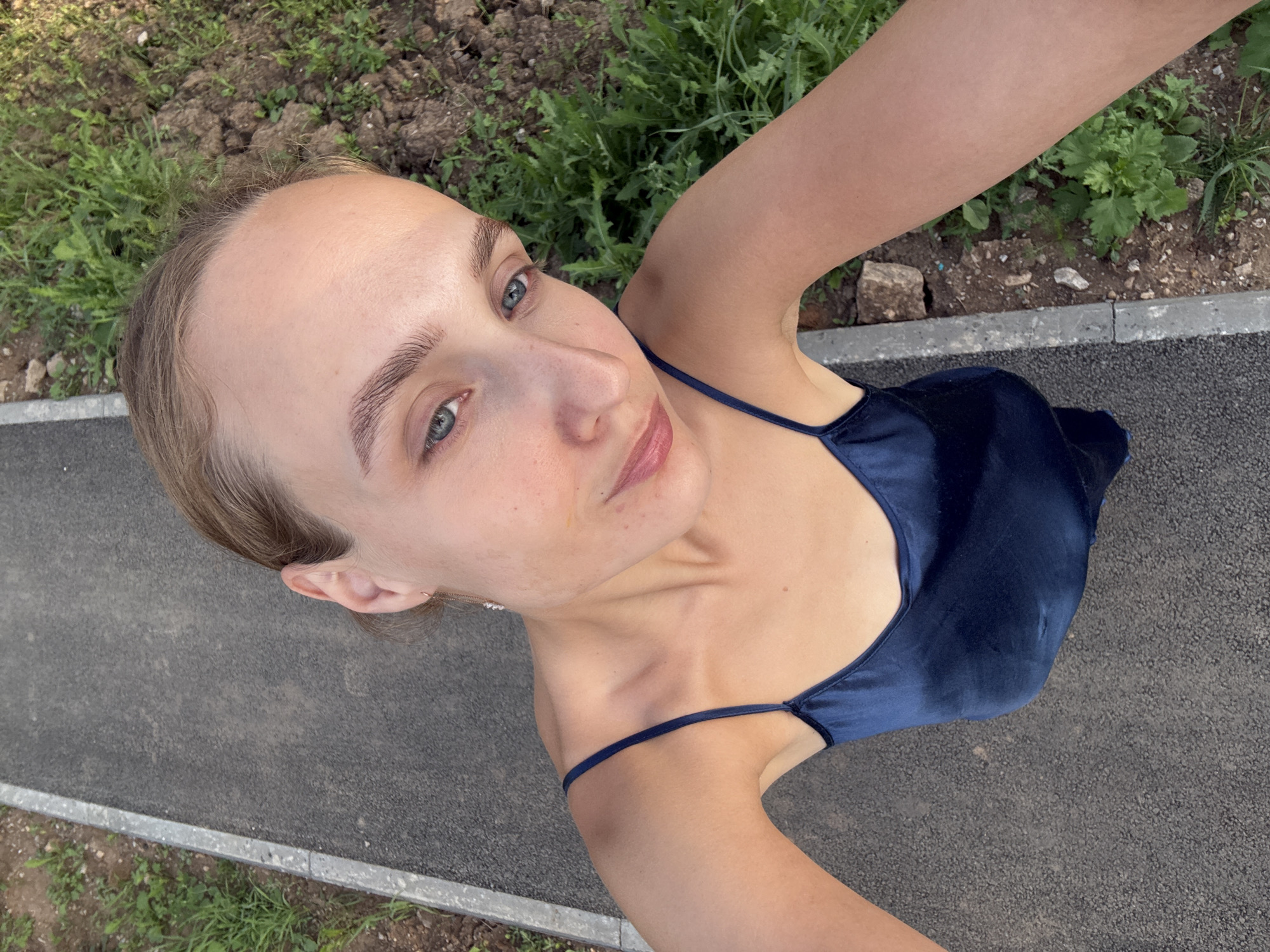 This media file features a young woman outdoors, captured from a high-angle perspective. She is looking upwards, with her face taking up a significant portion of the frame. She has light-colored hair, styled loosely, and her eyes are a striking blue-green. Her skin appears fair with some freckles visible on her nose and cheeks. She is wearing a dark navy blue satin dress with thin spaghetti straps. The background consists of a paved surface, likely a pathway or sidewalk, bordering a patch of green grass and soil. The lighting suggests it is daytime, with natural sunlight illuminating the scene. The woman's expression is neutral, perhaps a slight smile. The setting appears to be a park or a public outdoor area in Sharkan, Russia.