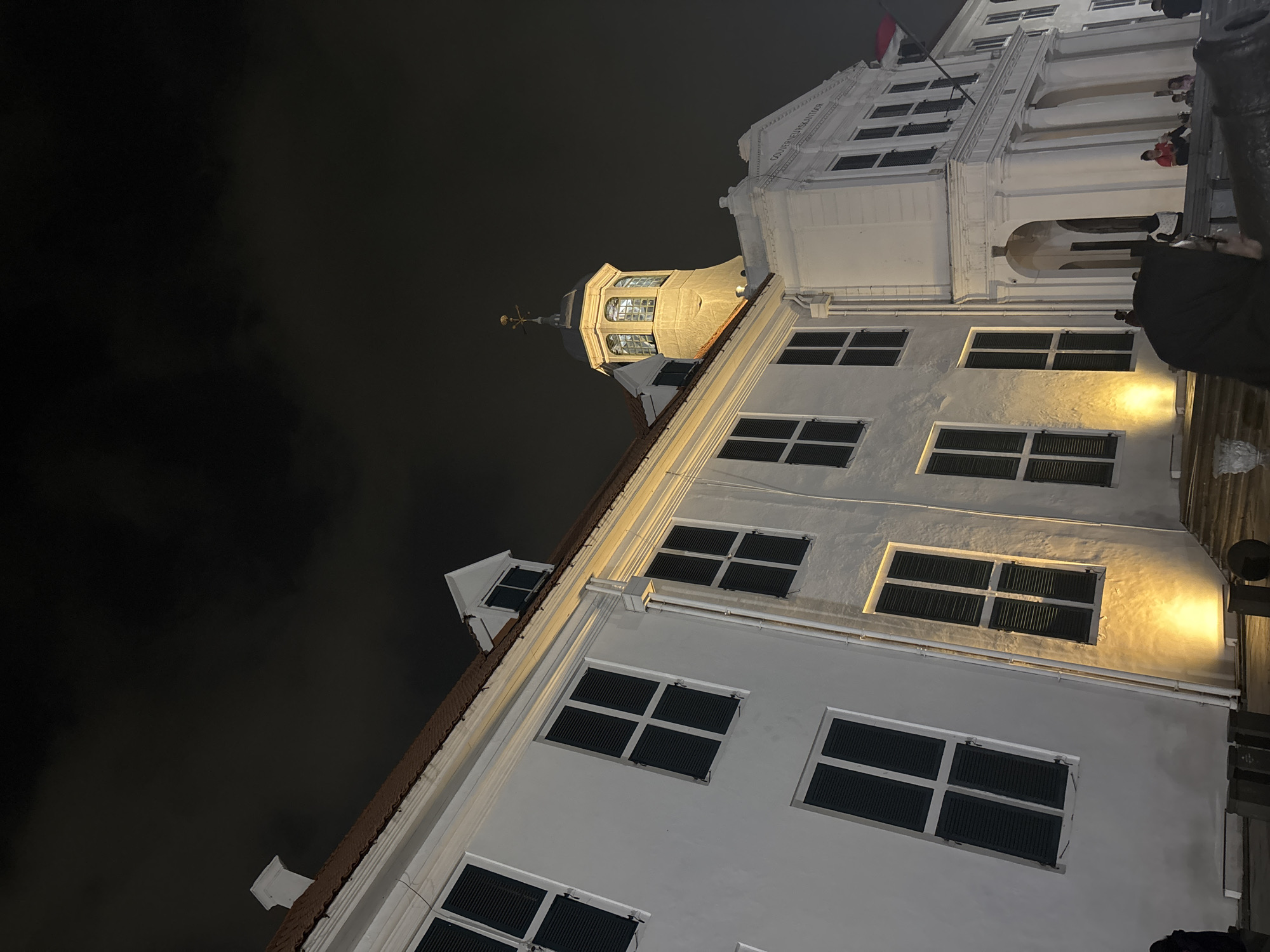 The image shows a night view of the Fatahillah Square in Kota Tua, West Jakarta, Indonesia. The main subject is the historic Dutch colonial building, the Jakarta History Museum (Museum Sejarah Jakarta), formerly known as the Old Batavia Town Hall. The building is illuminated by yellow floodlights, highlighting its white facade and numerous dark-shuttered windows. A prominent tower with a clock face and weather vane stands out against the dark, cloudy sky. In the foreground, there are some dark, indistinct objects that appear to be cannons. Several people are visible on the steps and in front of the building, some sitting and others standing, though their features are not clearly discernible due to the low light and distance. The overall impression is one of a historical landmark at night.