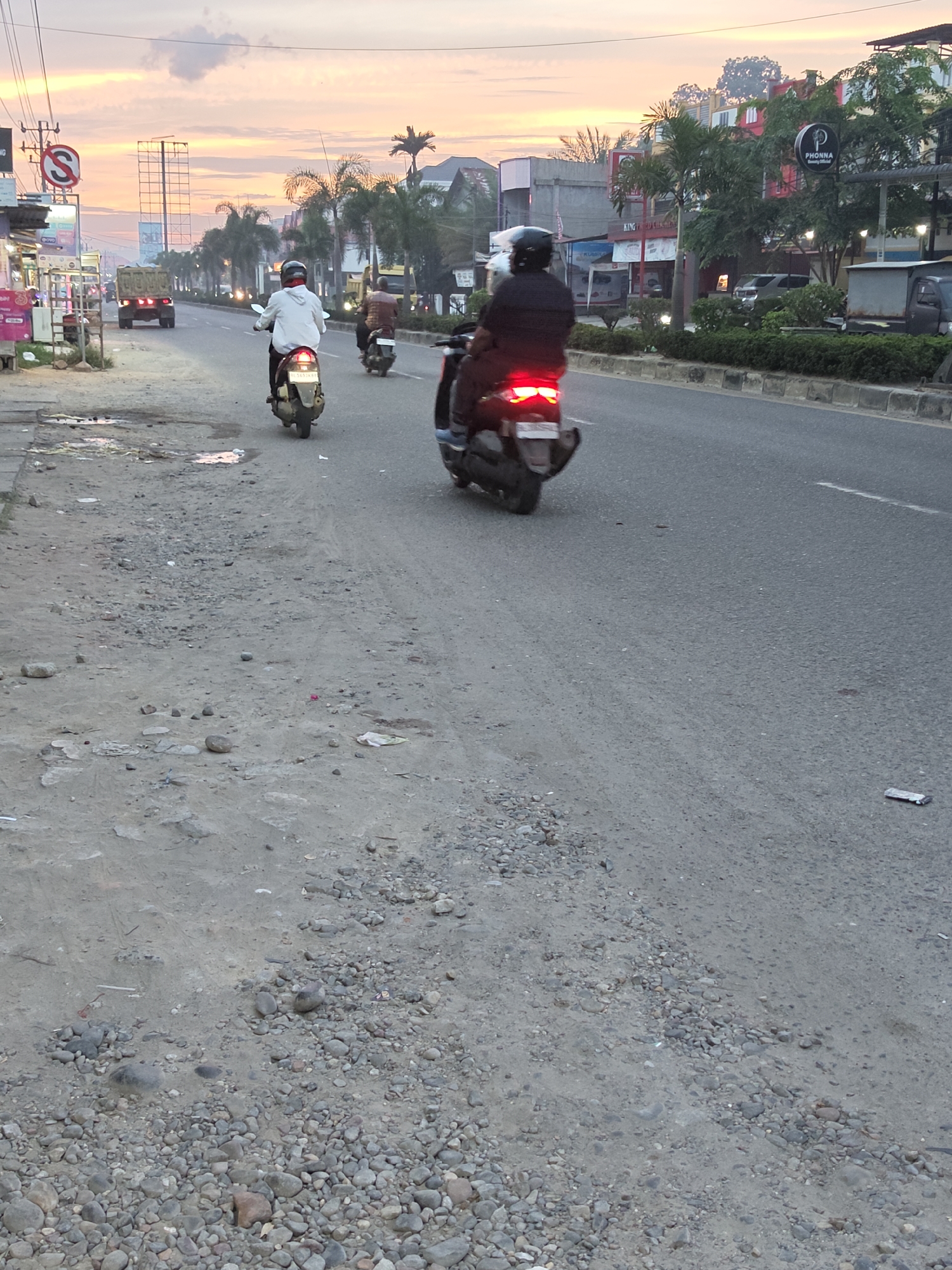The image depicts a street scene in Lhokseumawe, Indonesia, during what appears to be sunset. The sky is filled with warm, pastel colors, suggesting either sunrise or sunset. Several motorcycles are visible on the asphalt road, with riders wearing helmets. One motorcycle in the foreground has its brake lights illuminated. On the left side of the road, there are some buildings, shops, and a traffic sign indicating "S" with a diagonal line through it, likely a no-stopping or no-parking sign. A truck is also seen on the road further ahead. The roadside on the left appears to be a mixture of dirt, gravel, and debris. On the right side of the road, there are more buildings, some with lights on, and vegetation, including palm trees. The overall environment suggests an urban or suburban setting.