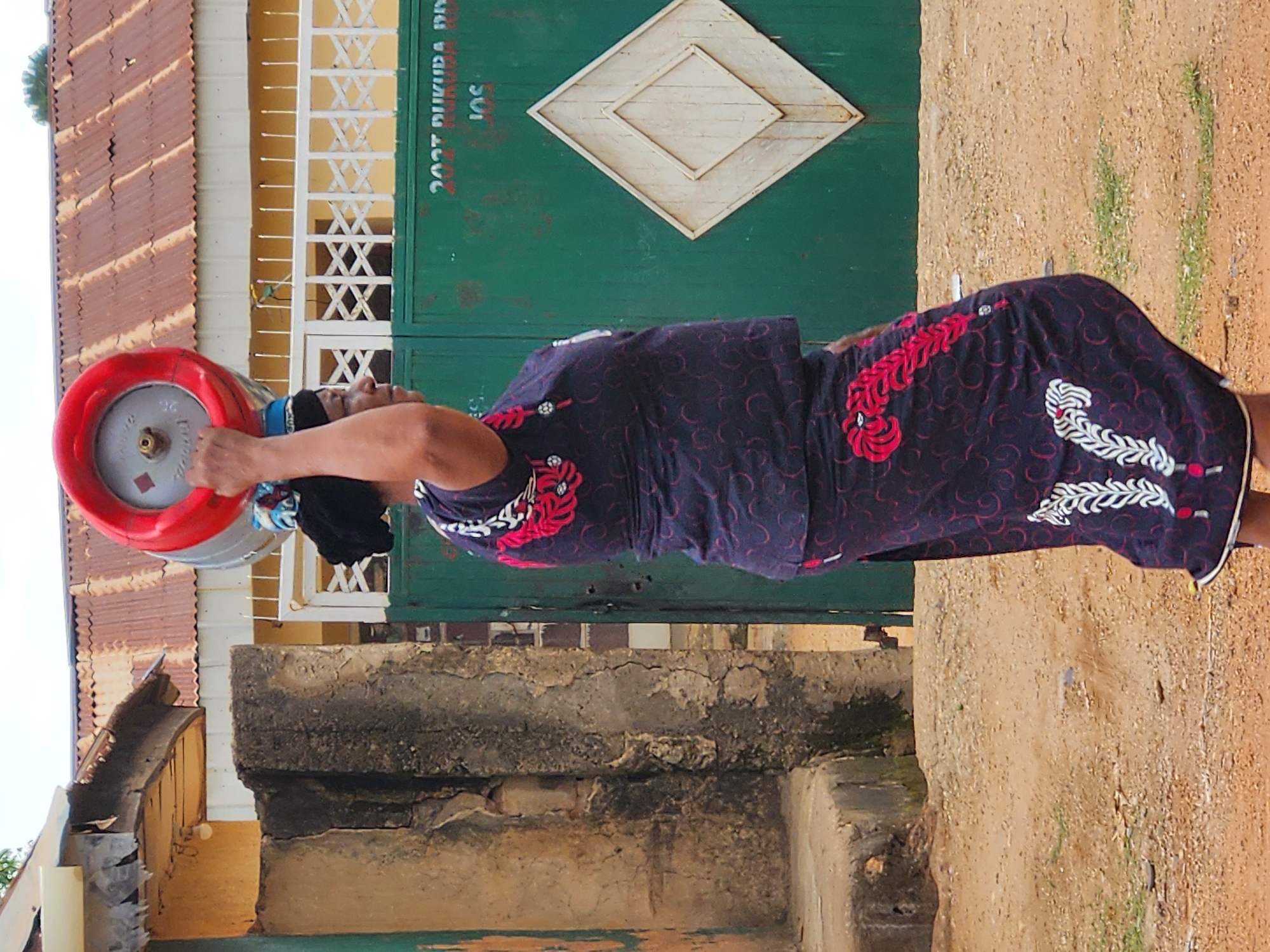 This image, oriented 90 degrees clockwise from its natural upright position, shows a woman carrying a gas cylinder in what appears to be an outdoor, semi-urban setting.
The main subject is a woman, seen from her right side, facing towards the left of the frame. She wears a dark blue or purple dress adorned with intricate red and white patterns, resembling feathers or leaves. Her hair is dark and pulled back, with parts of a blue and white head covering visible around her neck and ear. She is actively carrying a grey gas cylinder, identifiable as an LPG tank, which has a prominent red plastic cap or handle. She supports the cylinder with her right arm, resting it against her shoulder and head, indicating its weight.
The background features a large, dark green metal gate or door. In its upper center, a cream-colored, diamond-shaped decoration with a raised border is affixed. To the left of the gate, there's a light-colored building wall with several white grates, each forming an 'X' pattern. Above this wall, a weathered, corrugated metal roof, possibly rusty brown, is visible. The ground in the foreground and to the right is composed of light brown, rough dirt or sand, with sparse patches of green vegetation. Several layers of weathered concrete structures, possibly a low wall or foundation, run across the middle ground.
The activity depicted is the woman transporting the gas cylinder, suggesting a daily chore or errand. The bright, even lighting indicates it is daytime, likely clear and sunny.
Visible text includes "2023 DUKUDA" and "JOS" stenciled in white on the green gate. On the gas cylinder, faint text, possibly indicating "12KG" or "2KG" as a weight, is partially discernible.
