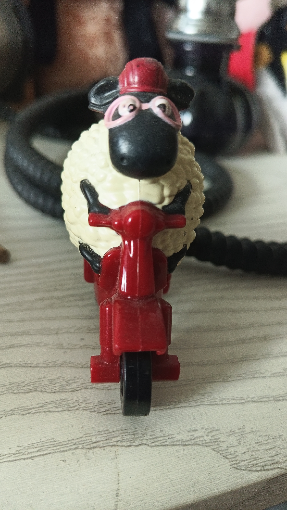 A close-up photograph shows a Shaun the Sheep toy figurine riding a red scooter. The sheep is wearing a red helmet and pink glasses. The background includes a black braided cord and a blurred object that appears to be part of a hookah. The surface beneath is a light-colored, wood-grained material. The photo suggests a playful or informal setting, as it features a child's toy placed on a wooden surface. The location details are unknown, as the image does not provide contextual information about the environment or city.