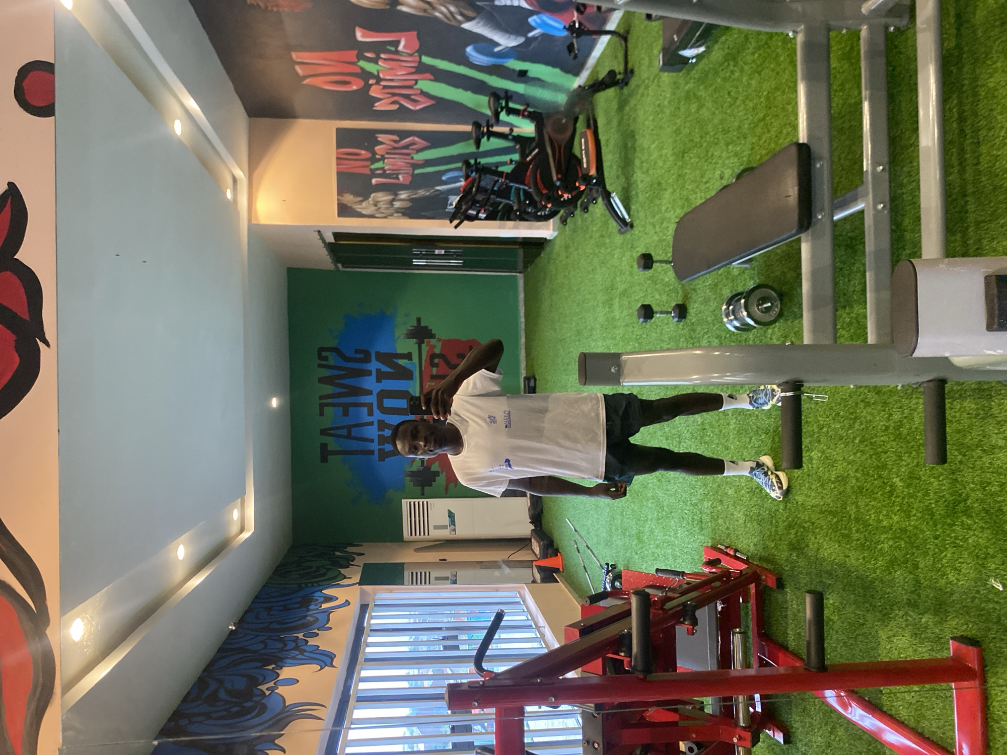 A man stands in a gym, taking a mirror selfie. He is wearing a white t-shirt and dark shorts, with white socks and patterned athletic shoes. The gym has bright, artificial lighting and is decorated with motivational murals on the walls. The floor is covered in green artificial turf, and various gym equipment is visible, including weight machines, exercise bikes, dumbbells, and a weight bench. To the left, a large red weight machine is prominent. In the background, there are two white air conditioning units and a window with blinds. The overall impression is of a modern, well-equipped fitness facility.