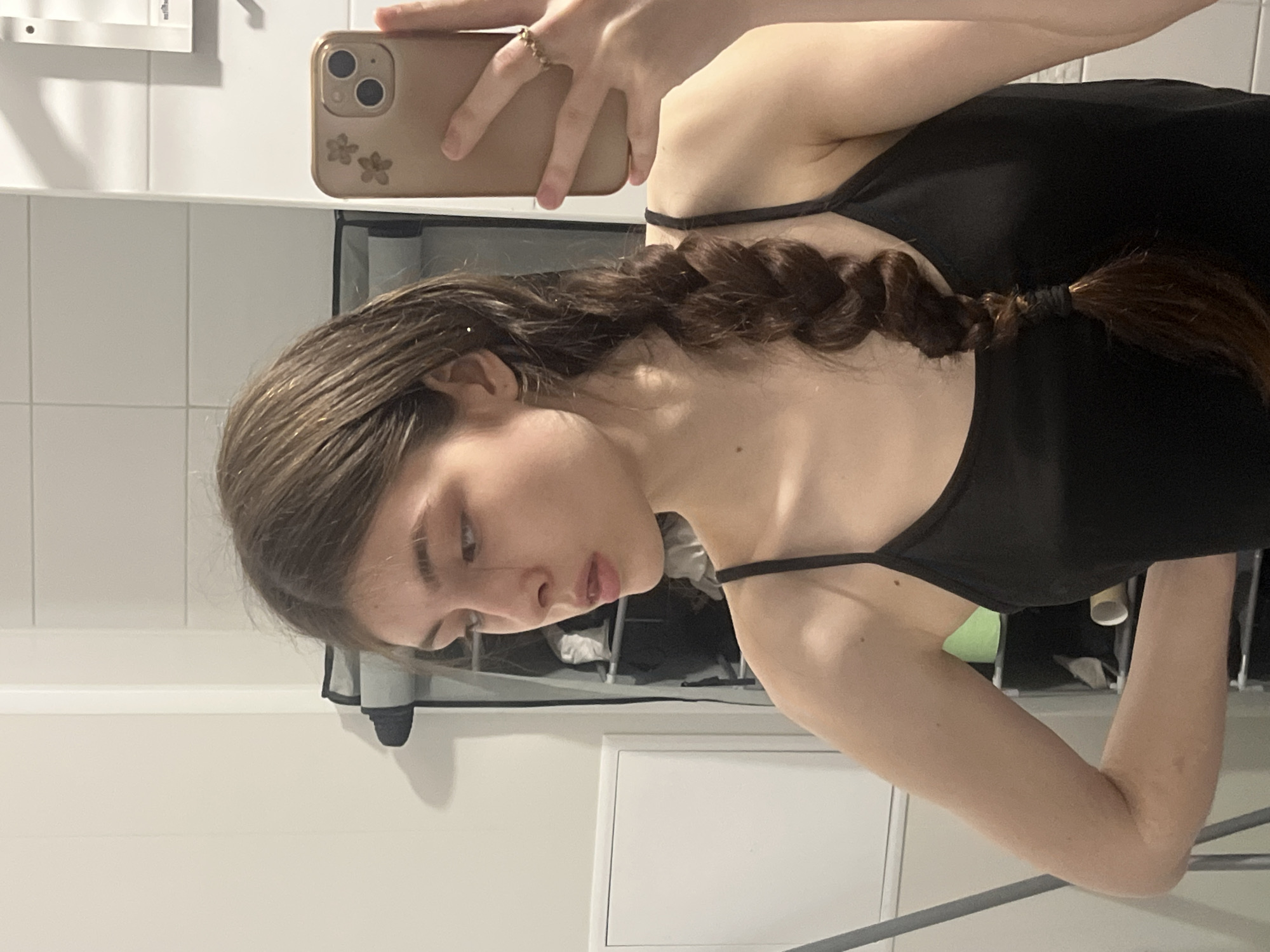 A young woman with brown hair braided to the side is taking a selfie in what appears to be a bathroom. She is wearing a black tank top. The background includes white tiled walls, a white cabinet, and a phone with a light brown case adorned with flower designs. The woman is holding the phone in her right hand, which also features a ring. The image's perspective is slightly tilted. The photograph does not contain enough details to determine the location within Saint Petersburg, Russia.