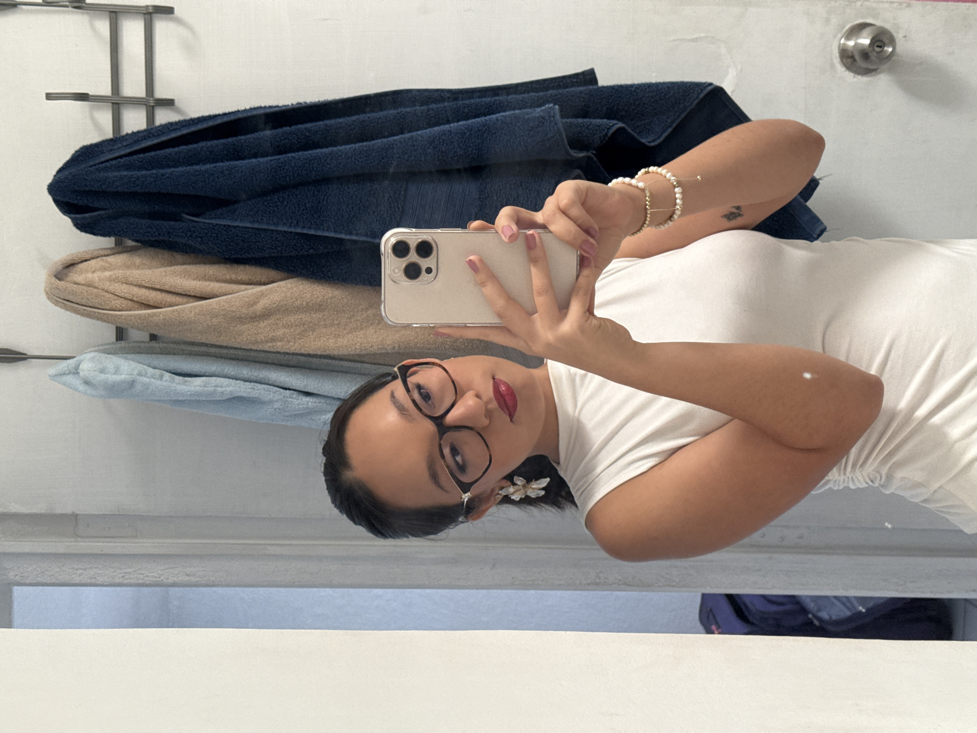 A woman with dark hair pulled back and glasses, wearing a white shirt and a flower earring, is taking a selfie with a beige-colored smartphone in a bathroom. She is positioned in front of a towel rack holding a dark blue towel, a tan towel, and a light blue towel. A pearl bracelet is visible on her wrist. A silver doorknob is visible on the wall above the towels. The bottom portion of the frame reveals a section of the white sink and a glimpse of a dark-colored object underneath. This setting is likely a residential bathroom, though without additional information, I cannot confirm the location of Puebla, Mexico.