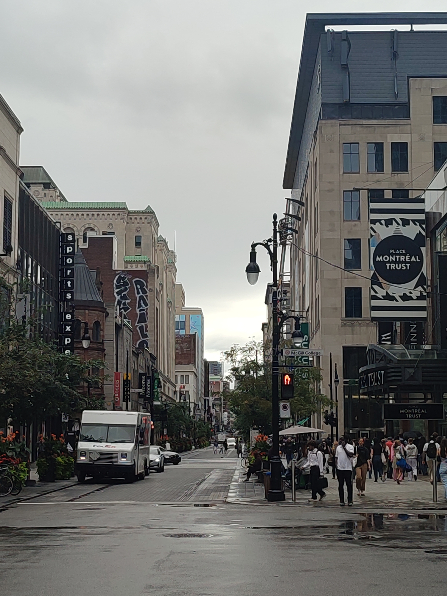 This image depicts a street scene in Montreal, Canada, likely on Sainte-Catherine Street, a well-known commercial artery. The day is overcast, and the wet pavement suggests recent rain.
On the right side of the street, a tall, modern building stands, adorned with large banners for "PLACE MONTRÉAL TRUST." The architecture features stone cladding and dark-framed windows. Below the banners, the entrance to the "MONTRÉAL TRUST" complex is visible. Pedestrians, some holding umbrellas, are walking along the sidewalk.
On the left side, older, more ornate buildings line the street, interspersed with modern glass facades. A vertical sign displays "sports exp" in black letters on a white background. Further down, graffiti is visible on a brick building. Trees and planters with orange flowers add greenery to the scene.
In the foreground, a white FedEx delivery truck is positioned in the street. The wet road surface reflects the surrounding buildings and lights. In the distance, the street stretches out, with more buildings and pedestrians visible, suggesting a busy urban environment. Traffic lights are visible, with one showing a red signal.