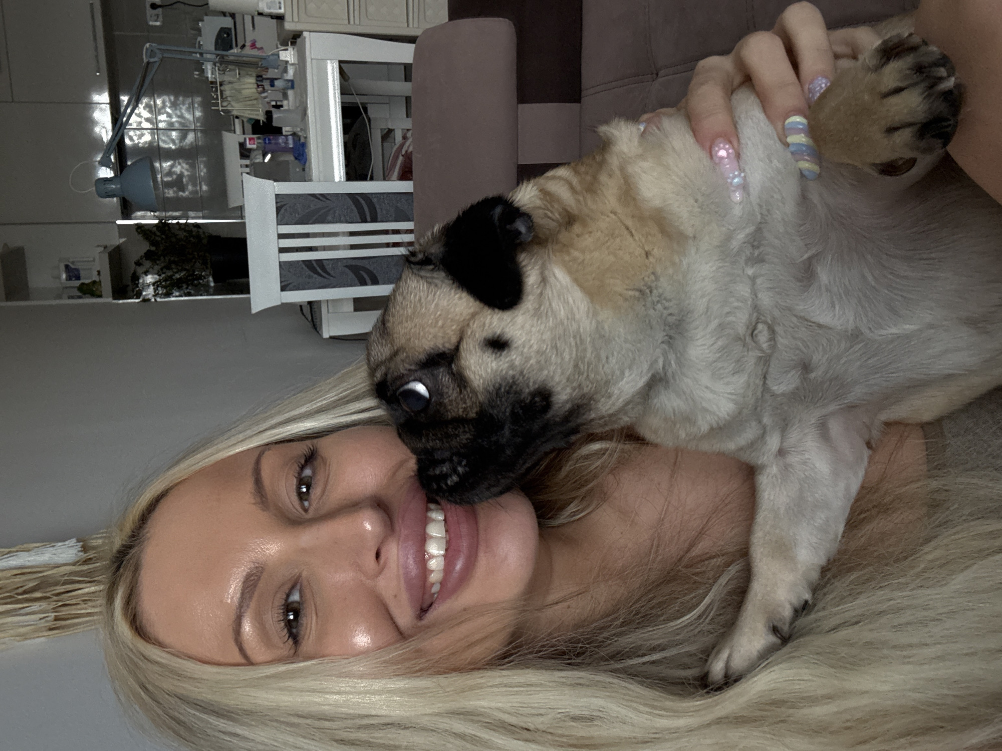 This is a medium shot, close-up, portrait-style photo that appears to be taken indoors. The primary subjects are a young woman with long blonde hair and a pug puppy. The woman is lying down, her head tilted back, and she is smiling widely. Her eyes are open and looking upwards. The pug puppy is positioned over her face, with its head nudging the woman's nose and mouth area. The puppy has its eyes wide open, looking towards the camera. The woman's hand, with long, colorful, decorated nails, is visible resting on the puppy. The background is blurred but shows what appears to be a domestic setting with furniture and what looks like a desk with various items, including a lamp, in the upper left corner. The lighting suggests it is daytime, likely indoors, as the light seems artificial and diffused. The mood is affectionate and playful, conveyed by the interaction between the woman and the puppy. There is no visible text in the image.