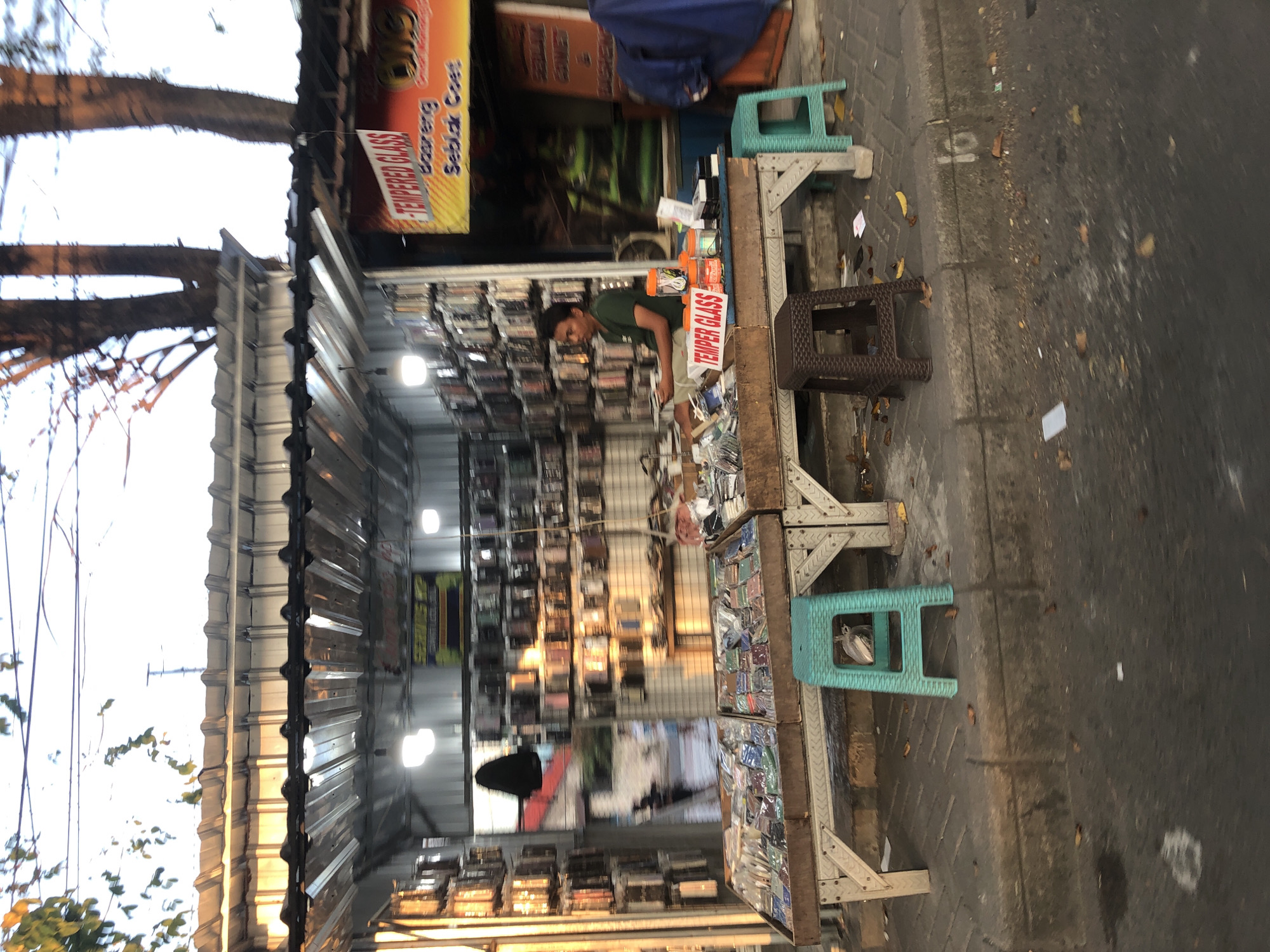 The image captures a street-side stall in Sepande, Indonesia, specializing in mobile phone accessories. The stall is covered by a corrugated metal roof supported by metal beams. The front of the stall displays an assortment of phone cases and tempered glass screen protectors laid out on two tables. Behind the tables, shelves are packed with more phone cases. A young man, wearing a green t-shirt and shorts, is seated at the stall, appearing to be the proprietor or an employee. He is looking towards the left of the frame. Several plastic stools, in green and brown, are placed around the stall. The street in front of the stall is paved with interlocking bricks, and a small bird is perched on one of the green stools. The background shows a street with some trees and a hazy sky, suggesting it might be early morning or late afternoon. A prominent orange banner with the words "TEMPERED GLASS" is visible on the right side of the stall, indicating the primary product offered.