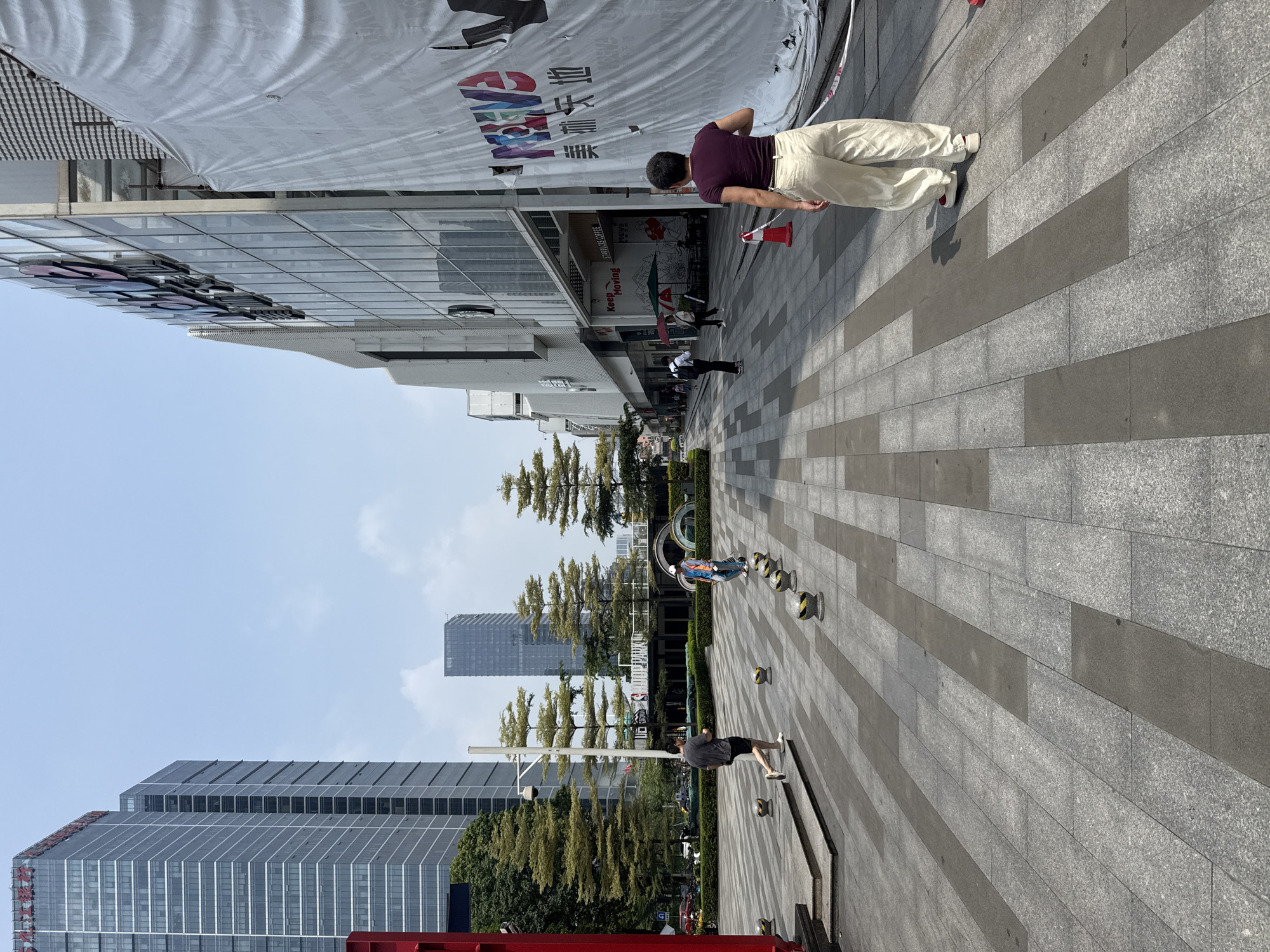 A street scene in Tianhe District, China, shows a wide, paved pedestrian walkway lined with modern buildings and trees under a clear sky. In the foreground, a woman with short dark hair wearing a purple t-shirt and loose cream-colored pants stands with her back to the camera, facing away from the viewer and towards the buildings. To her left, a man in a dark t-shirt and shorts walks away from the camera. Further down the walkway, another person in a bright orange and blue vest is walking. The buildings on the right are a mix of glass-fronted structures, with one large building featuring a white banner with colorful lettering. On the left, tall, dark skyscrapers with reflective windows dominate the background, interspersed with lush green trees. Several small, round, yellow and black bollards are visible along the walkway. Traffic is visible in the distance on the left.