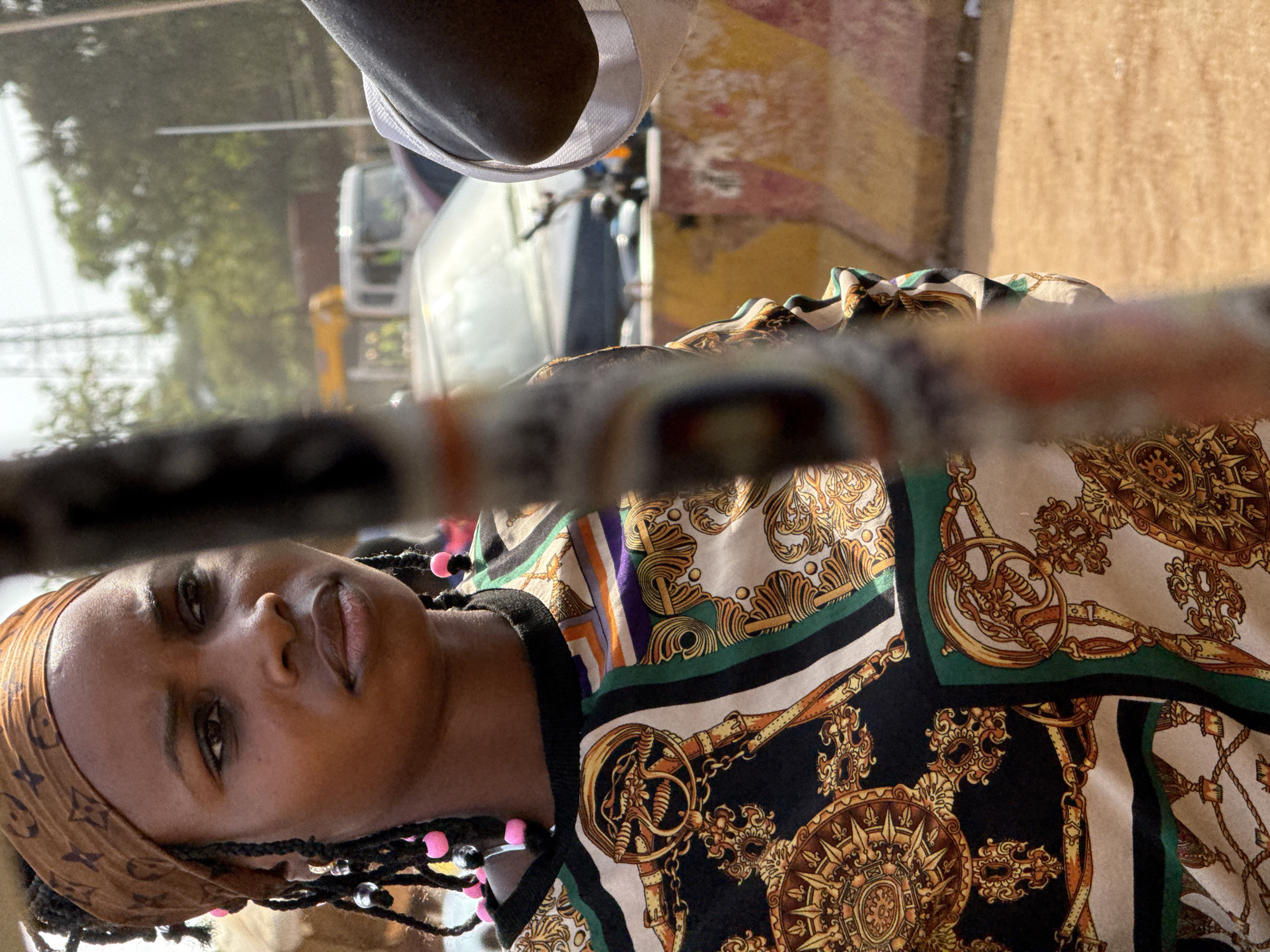 The image shows a woman with dark skin, a brown Louis Vuitton patterned head wrap, and dark braids adorned with pink and silver beads. She is wearing a black, cream, and gold patterned blouse. The photo appears to be taken from inside a vehicle, with a portion of the steering wheel visible in the foreground. Through the car window, there is a glimpse of a white vehicle and a building with a light brown exterior. Given the architecture and the woman's attire, it is plausible this image was taken in Abuja, Nigeria.