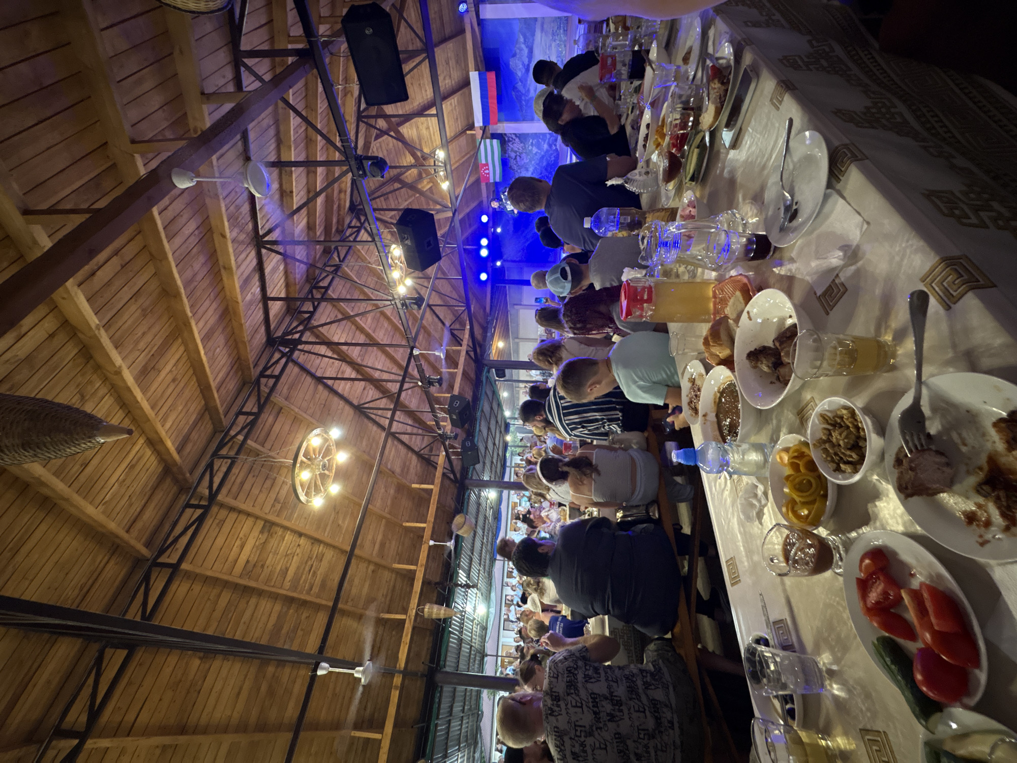 The image depicts an indoor gathering of people at long tables laden with food and drinks, possibly a restaurant or event hall, located in Durypsh, Abkhazia. The setting features a wooden ceiling with exposed beams and industrial-style lighting fixtures, including chandeliers and ceiling fans. A stage with a performer and a backdrop displaying mountainous scenery and national flags of Abkhazia and Russia is visible in the background, illuminated by blue lights. Many individuals are seated and engaged in eating or conversing. On the tables, plates with food, bowls of various dishes, glasses, and bottles are arranged. The overall atmosphere suggests a lively social event or a festive meal.