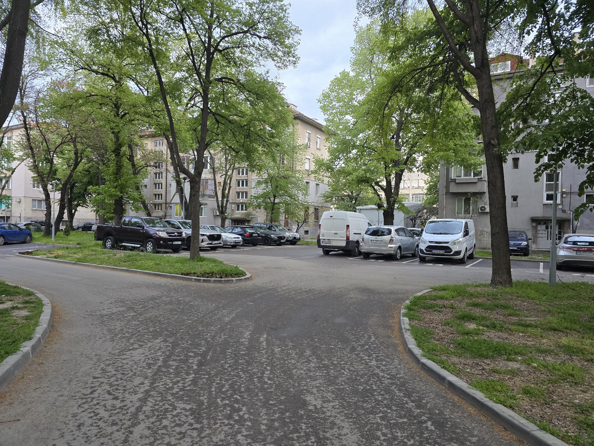 This image depicts a quiet, tree-lined urban street and parking area in Plovdiv, Bulgaria, during daytime on an overcast day. No people are visible.
The scene is dominated by a paved asphalt road that forks, leading to multiple cars parked on both sides. The road surface shows signs of wear and patchy discoloration. Concrete curbs separate the asphalt from narrow strips of sparse grass and earth, where tall deciduous trees with vibrant green leaves stand, suggesting spring or early summer.
Numerous vehicles are parked: on the left, a blue sedan, a dark brown Toyota Hilux pickup truck (with Bulgarian license plate `PB 0205 KA`, indicating Plovdiv), a silver Mazda CX-5, and other cars are visible. On the right, a white Ford Transit van, a silver Toyota Yaris (`CB 2911 MA` - Sofia), another white Ford Transit van (`CH 9036 KB` - Haskovo), a silver Subaru Impreza (`PB 3553 BT` - Plovdiv), and a dark blue Volkswagen Golf (`A 9703 MC` - Burgas) are parked.
In the background, multi-story residential buildings with light-colored (beige and grey) facades and multiple windows are partially visible through the dense tree canopy. One building on the right has a partial number "71" on its facade. The overcast sky provides soft, diffused lighting, eliminating harsh shadows. The overall impression is a typical, tranquil residential street scene with ample greenery and parked cars.