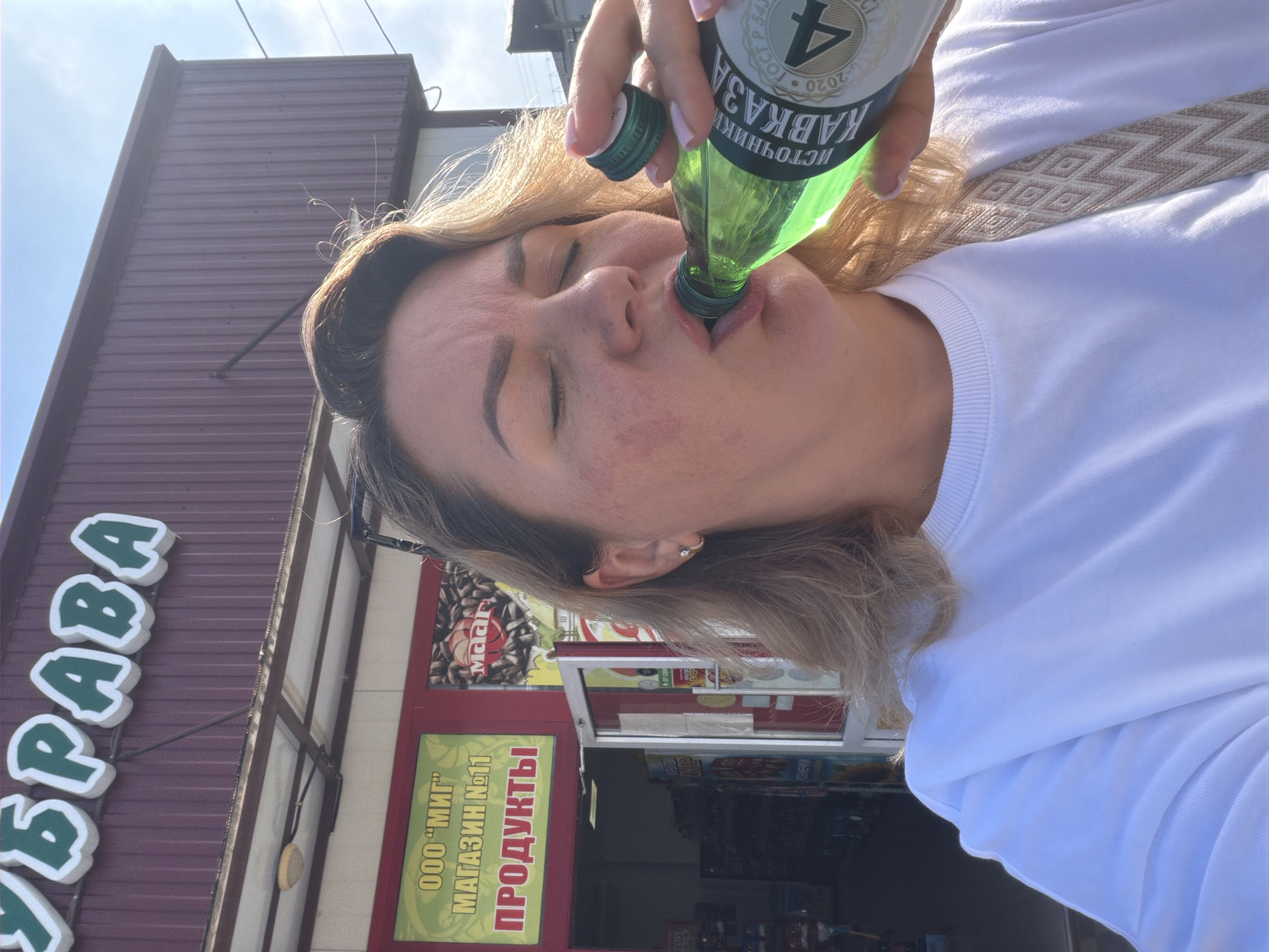 A woman with blonde hair is drinking from a green glass bottle of mineral water in front of a store. The store has a red facade and a sign that says "УБРАВА" in large white letters with a green outline. Below this sign, there is a smaller sign in Russian that says "ООО "МИГ" МАГАЗИН №11 ПРОДУКТЫ", indicating it is a store named "MIG" and sells groceries. The bottle has a label that reads "ИСТОЧНИКИ КАВКАЗА" and a number "4". The woman is wearing a white sweatshirt and has a strap with a geometric pattern across her chest. The scene appears to be outdoors on a sunny day. Leningradskaya, Russia is a town in Krasnodar Krai, but there is no specific visual indicator to confirm the location beyond the text on the signs.