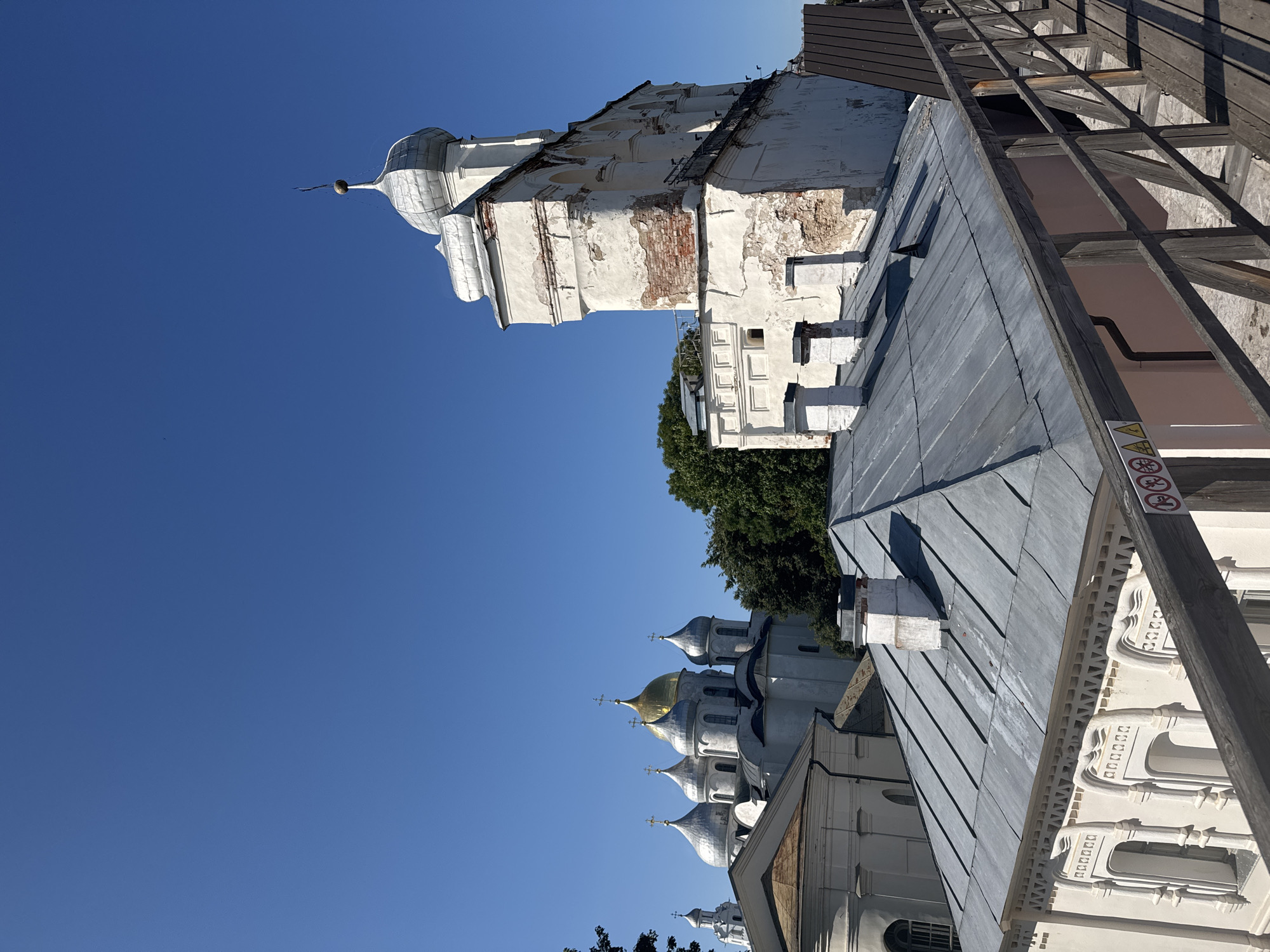 The image depicts the architectural ensemble of the Veliky Novgorod Kremlin, also known as Detinets, under a clear blue sky. In the foreground, a weathered wooden walkway with a railing overlooks several rooftops. The rooftops are constructed of grey metal and feature several white chimneys.

Dominating the mid-ground and background are various religious structures characteristic of Russian Orthodox architecture. To the left, a cluster of cathedrals with silver domes, one of which is gilded, are visible. These buildings are predominantly white with arched windows. To the right of these cathedrals, a tall, white bell tower with visible signs of aging, including crumbling plaster and exposed brickwork, rises against the sky. This tower is topped with a silver dome. Mature green trees are interspersed between the buildings. The overall scene conveys a sense of historical significance and architectural grandeur.