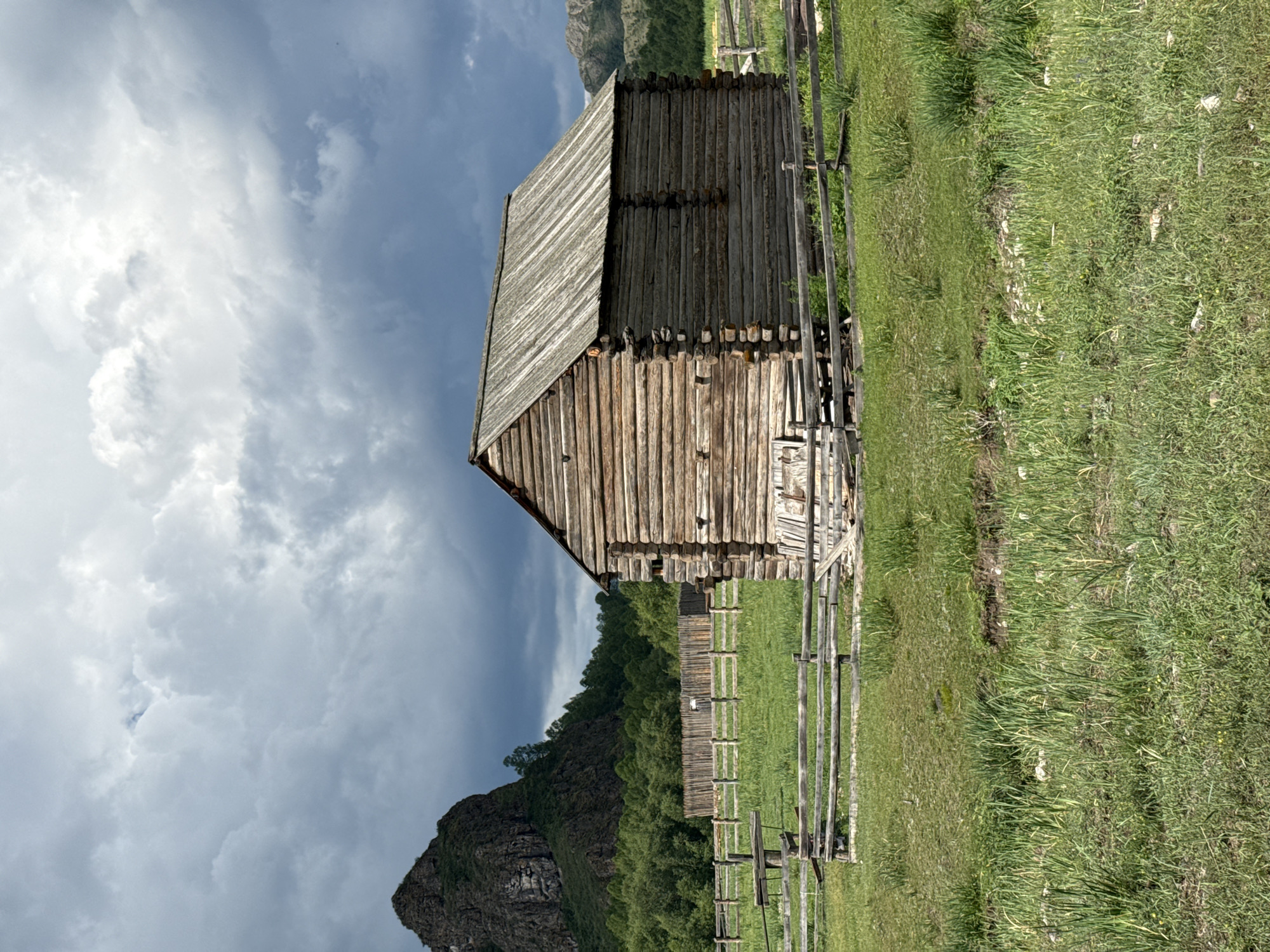 The image depicts a rustic wooden structure, likely a barn or storage building, situated in a rural landscape. The building is constructed from rough-hewn logs, with a weathered wooden roof. It is enclosed by a simple wooden fence that demarcates a grassy area. In the background, a large, rocky mountain peak rises against a dramatic, cloudy sky. Dense green foliage, including trees, forms a band between the fenced area and the mountain. Further in the distance, more rolling, forested hills are visible. The foreground consists of a field of tall, green grass. The overall scene suggests an outdoor, natural environment, possibly a farm or a remote settlement, in the area of Kazanovka, Russia. There are no people visible in the image.