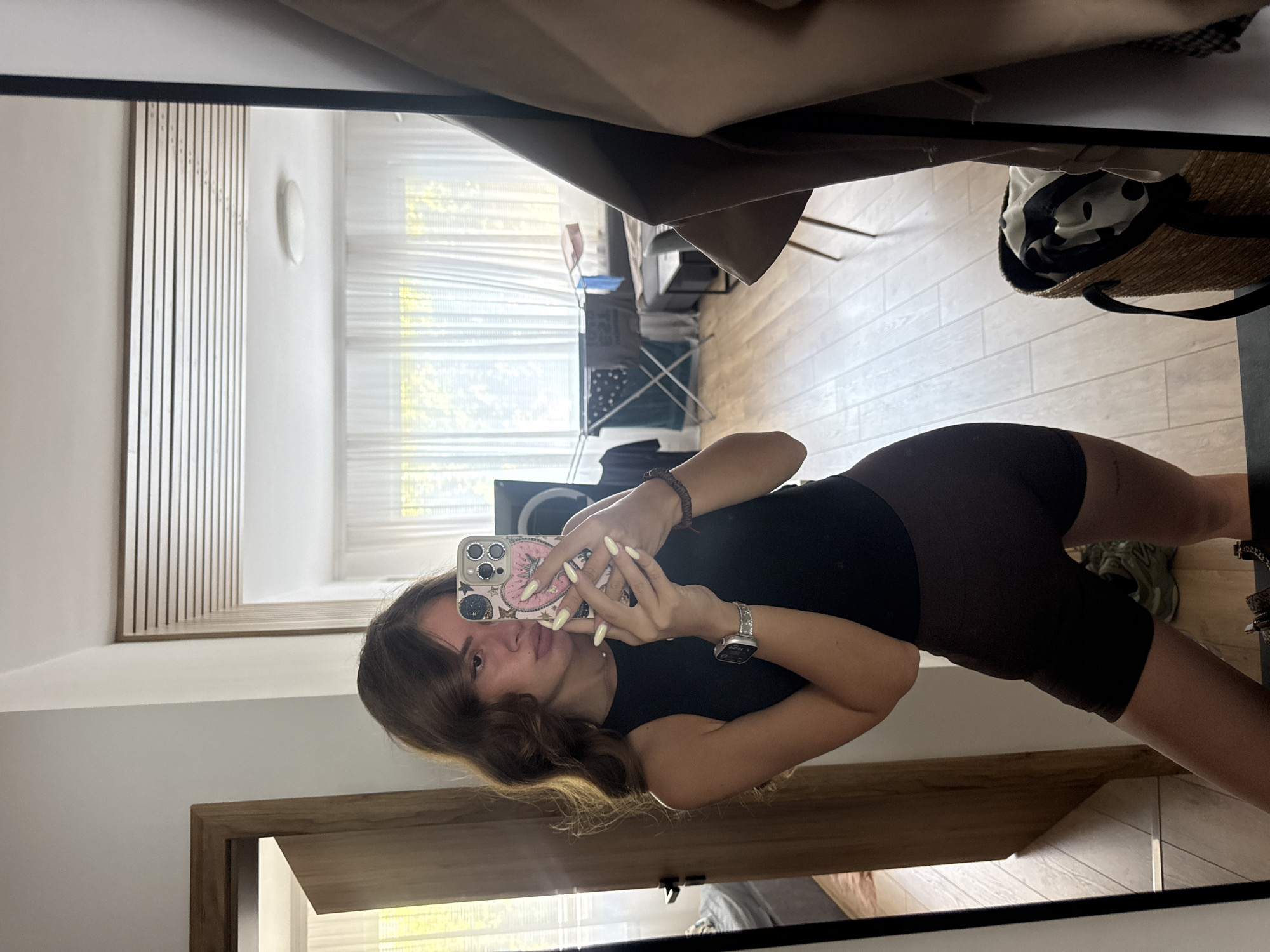 A young woman with wavy brown hair is taking a selfie in a mirror. She is wearing a black tank top and brown athletic shorts. She has long, light-colored fingernails and is wearing a silver watch on her left wrist. Her phone, which has a pink case with heart and star designs, is held up to take the picture. The reflection shows a room with light-colored walls and wooden floors. In the background, there are sheer white curtains, a drying rack with clothes, and a glimpse of a doorway with a bed visible beyond it. To the right of the woman, a tan coat is draped over something, and a woven basket with black and white patterned fabric is on the floor. The overall scene appears to be a domestic interior, likely a home. There is no indication of the city of Plovdiv, Bulgaria.