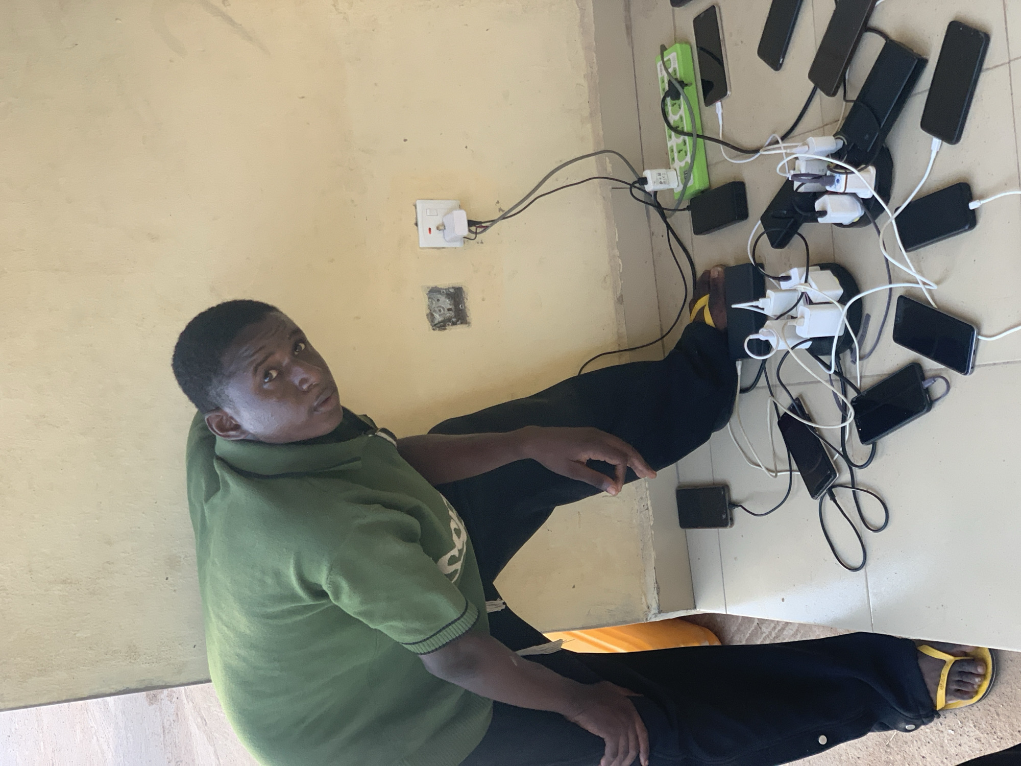 An indoor scene in Lokoja, Nigeria, shows a man sitting on a tiled floor surrounded by numerous mobile phones being charged. The man, wearing a green polo shirt, dark trousers, and yellow flip-flops, is seated with his legs bent and his body angled towards the viewer, making direct eye contact with a neutral expression.
To his right, on the light-colored tiled floor, are approximately 10-15 black mobile phones, all connected via charging cables to power adapters. These adapters are plugged into at least two power strips: one green, labeled "E-CHARGE," and another black, multi-outlet circular/oval strip. The cables create a dense network across the floor, highlighting a concentrated charging activity. A functional white wall socket, with an adapter plugged in, is visible on the light yellow wall, alongside a lower, empty or damaged socket recess. The lighting appears bright and even, suggesting daytime. The setup indicates a significant demand for phone charging, potentially in a communal setting or a place with limited access to power outlets.