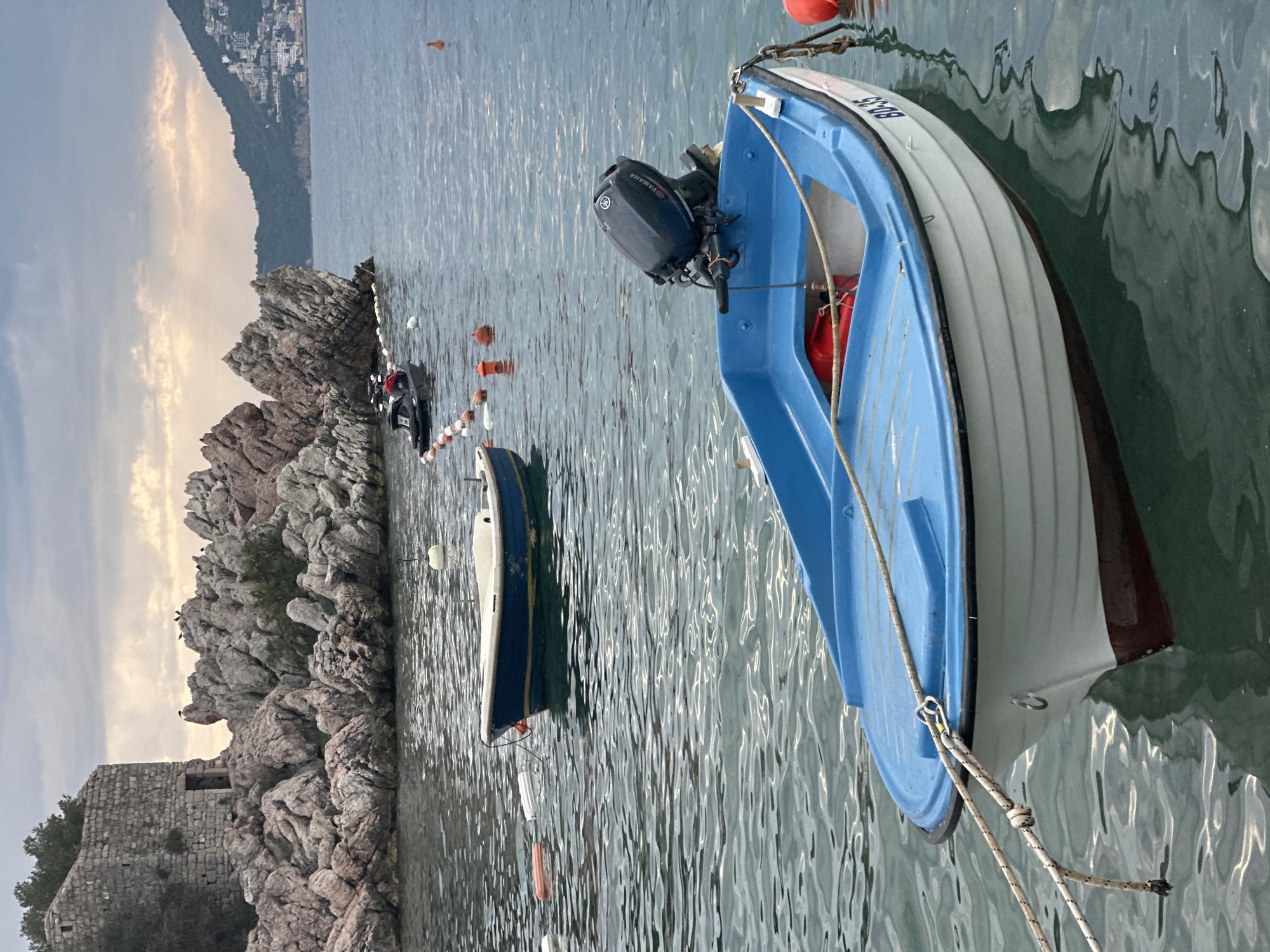 In the coastal town of Pržno, Montenegro, a serene scene unfolds on a body of water. In the foreground, a small, blue and white motorboat with a Yamaha outboard motor is tethered by ropes, its hull reflecting in the gentle ripples. Further out, another boat, blue and white with a yellow stripe, floats serenely. A black jet ski is visible in the middle distance, anchored near a line of orange buoys. The background is dominated by a rugged, rocky outcrop crowned with a weathered stone structure, possibly an old watchtower or fort. On the rocks, several birds are perched. Beyond the rocks, the coastline stretches out, featuring a lush green hillside dotted with buildings, suggesting a larger settlement. The sky is overcast with a hint of soft, diffused light.