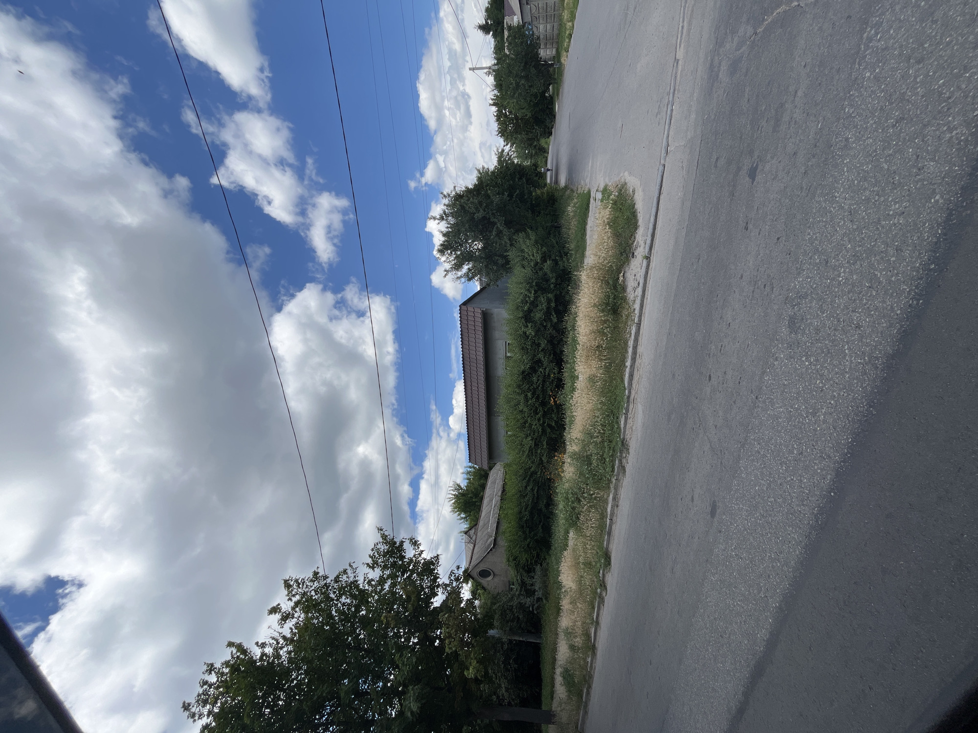 This is a vertical, low-angle outdoor shot of a street in Dnipro, Ukraine. The sky is a vibrant blue, dotted with fluffy white clouds and crisscrossed by power lines. To the left of the frame, a dense green tree grows tall against the sky. In the middle ground, a low-slung building with a brown tiled roof is partially visible behind a neatly trimmed hedge. Further back, another structure with a sloped roof and a round window stands amidst lush greenery. The right side of the image is dominated by a grey asphalt road, bordered by a strip of dry, light-brown grass and a thin concrete curb. The overall scene suggests a sunny day, possibly late morning or early afternoon, in a semi-rural or suburban setting. No people are visible in the frame.