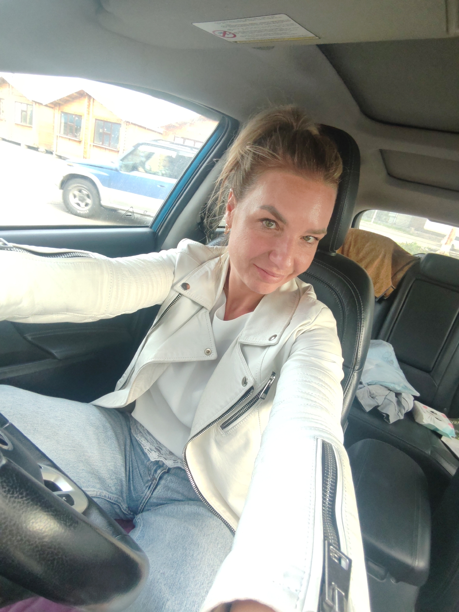 The image shows a woman in the driver's seat of a car, taking a selfie. She is wearing a white leather jacket and blue jeans. Her hair is pulled back in a ponytail. The interior of the car is visible, including the steering wheel, dashboard, and seats. Through the car window, there are buildings and a blue vehicle parked outside. The scene appears to be in a residential area. The lighting suggests it is daytime.