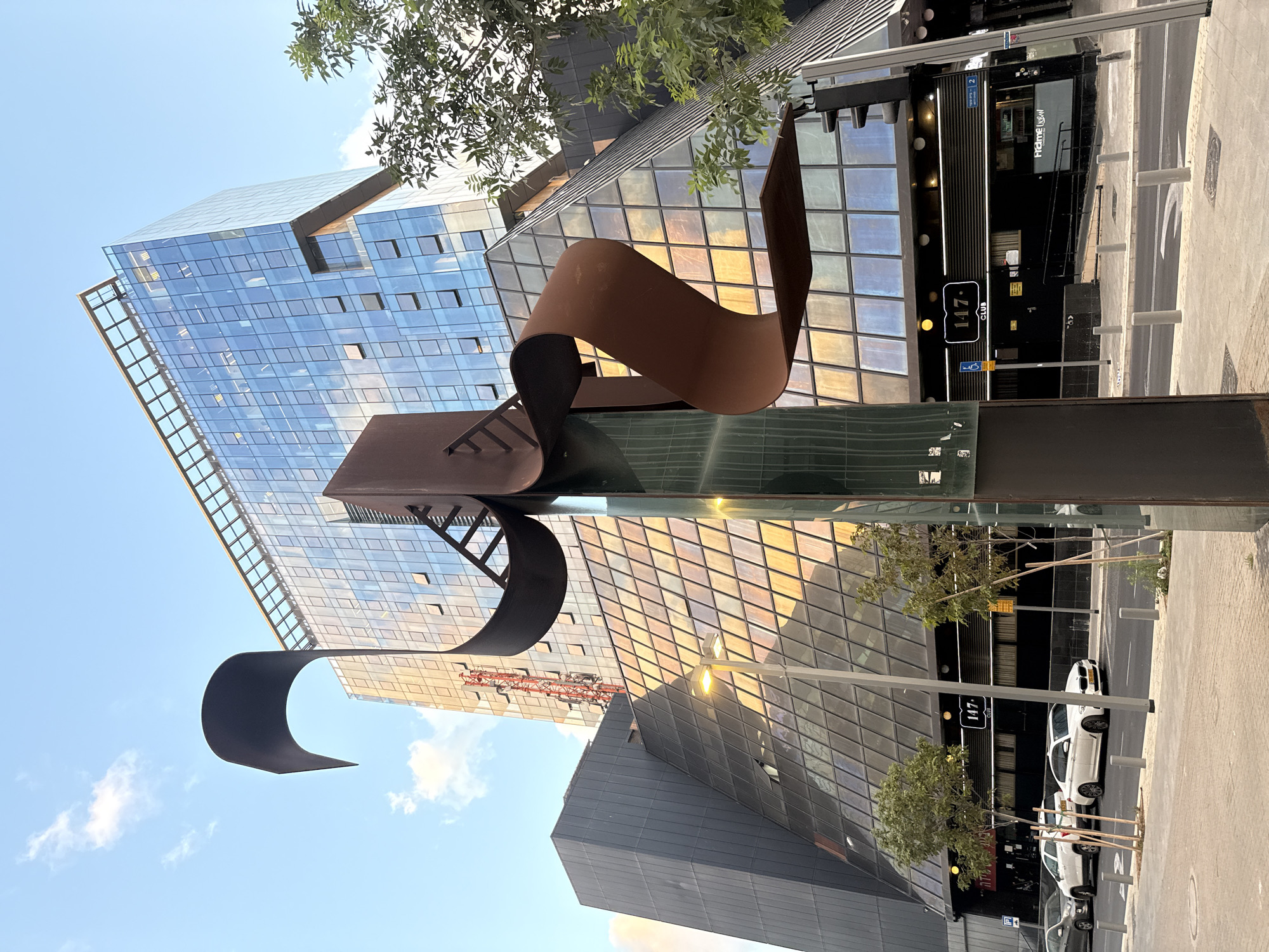 The image depicts a vibrant urban scene in Tel-Aviv, Israel, showcasing modern architecture and public art under a clear blue sky. A prominent, multi-story building with a facade of reflective glass dominates the background. Its numerous square and rectangular windows mirror the sky and warm, golden light, suggesting late afternoon or early morning. A red construction crane is visible as a reflection on the building's surface.
In the foreground, an abstract sculpture made of weathered, rust-colored metal stands as a striking art installation. Its curvilinear forms interlock and extend upwards, creating a dynamic shape against the backdrop of the glass building.
To the right, another building features commercial establishments at street level. Visible signs include "147° club" and "Framer Crew." The Hebrew text "שדרות רוטשילד 2" (Rothschild Blvd 2) is also displayed, along with a wheelchair accessibility symbol. The street area includes a paved sidewalk, gray bollards, and two white cars parked along the curb. A city bus, partially visible on the right, displays the number "117." Green foliage from scattered trees adds a natural touch to the otherwise architectural setting. The bright lighting and reflections convey a sunny, clear day.