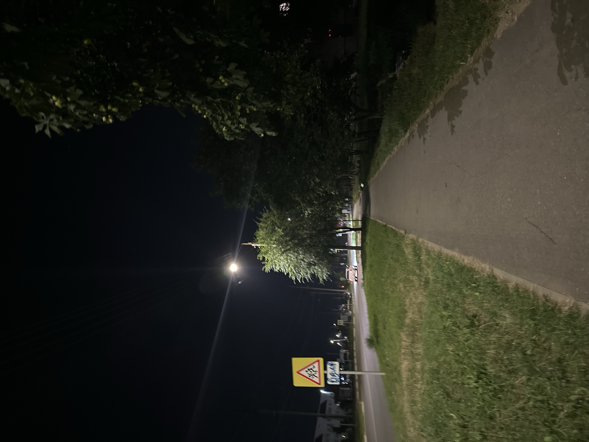 The image depicts a nighttime scene on a street in Nevinnomyssk, Russia. A paved sidewalk runs along the right side of the frame, bordered by a grassy area and trees. A street lamp illuminates the scene, casting a warm glow on the sidewalk and the surrounding vegetation. Across the road, which appears to be a multi-lane street, there are buildings and more trees. A yellow diamond-shaped road sign with a red border and a black symbol indicating a pedestrian crossing is visible on the left side of the road. A small, red car is seen driving on the road in the distance, its headlights on. The sky is dark, indicating nighttime, and the overall atmosphere is quiet and peaceful.