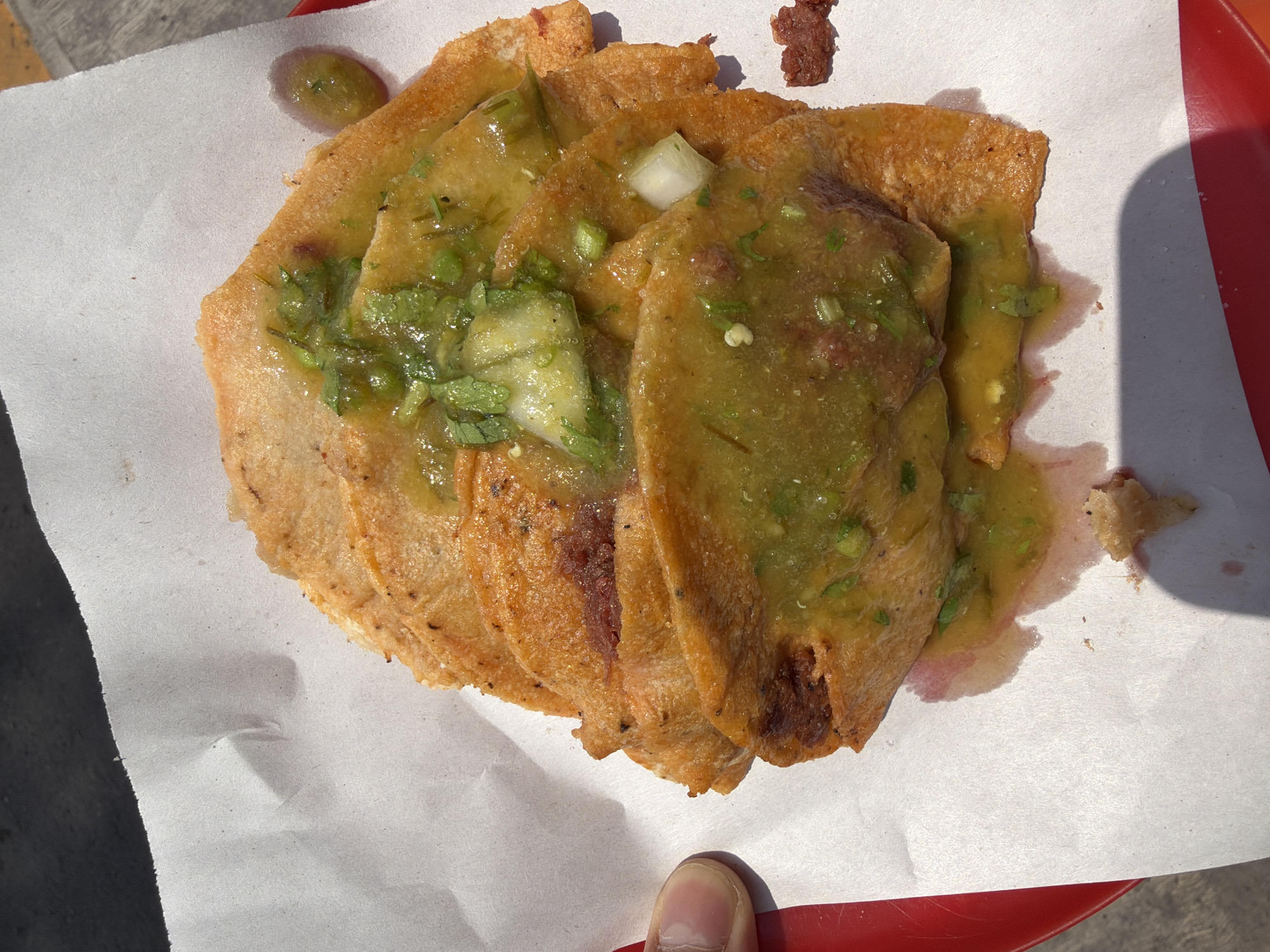 The image shows a plate of golden-brown tacos dorados (crispy tacos) covered in green salsa. The tacos are arranged on a white paper liner, which rests on a red tray. A fingertip is visible at the bottom of the frame, near the tacos. The photograph was likely taken in Puebla, Mexico, as tacos dorados are a regional specialty. The tacos appear to be freshly prepared and served.