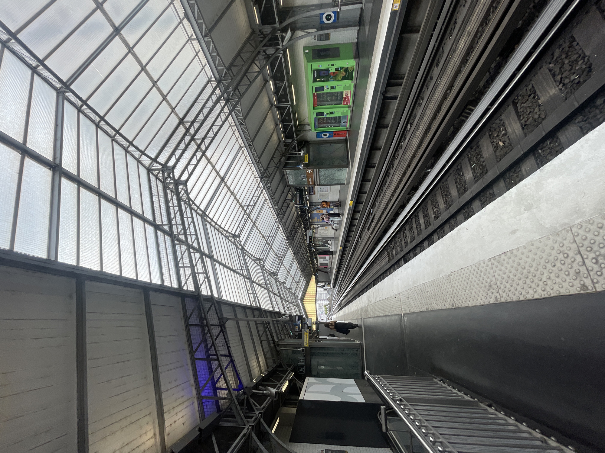 The image shows a view from a metro station platform in Paris, France, likely an elevated or open-air station due to the large, transparent roof structure. The roof is supported by a metal framework and covered with translucent panels, allowing natural light to filter through. On the left, a person in dark clothing walks away from the camera on a black platform. A metal railing separates the platform from the edge. To the right of the tracks, there is another platform with various elements including a bright green vending machine, signage, and a circular blue and white sign. Several people are visible on this platform. The metro tracks run along the center of the image, with ballast and sleepers clearly visible. In the distance, the city skyline can be seen through the open end of the station, with some buildings and trees. The overall impression is one of an active urban transportation hub.