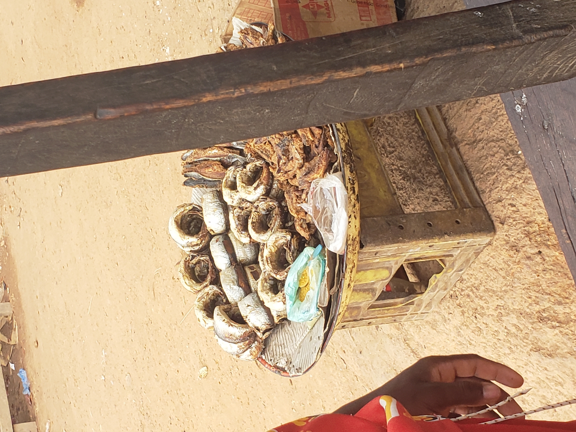 A market stall in Gumel, Nigeria, displays a variety of dried fish for sale. The fish are arranged on a circular tray, which rests on a yellow plastic crate. Some fish are whole and ring-shaped, while others are broken into smaller pieces. A few small plastic bags containing what appears to be spices or seasoning are placed among the fish. In the foreground, the arm and hand of a person wearing a red garment are visible on the left side of the frame. The ground is sandy and dry. A thick, dark wooden post stands vertically on the right, partially obscuring the view of the stall. In the background, there are hints of more market activity and debris on the ground.