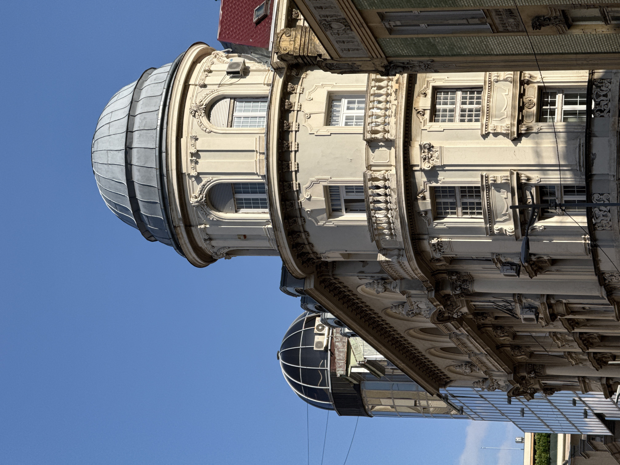 The image shows the facade of the Hotel Moskva in Belgrade, Serbia. The hotel is a landmark building located at the intersection of Terazije and Balkanska streets. The image focuses on the upper floors of the hotel, particularly a distinctive curved corner with a dome. The building exhibits architectural details typical of Art Nouveau and Secessionist styles, with ornate decorations, arched windows, and a curved balcony. A modern glass building is visible to the left of the Hotel Moskva, creating a contrast between old and new architecture. The sky is clear and blue, indicating a sunny day. No people are visible in the image.