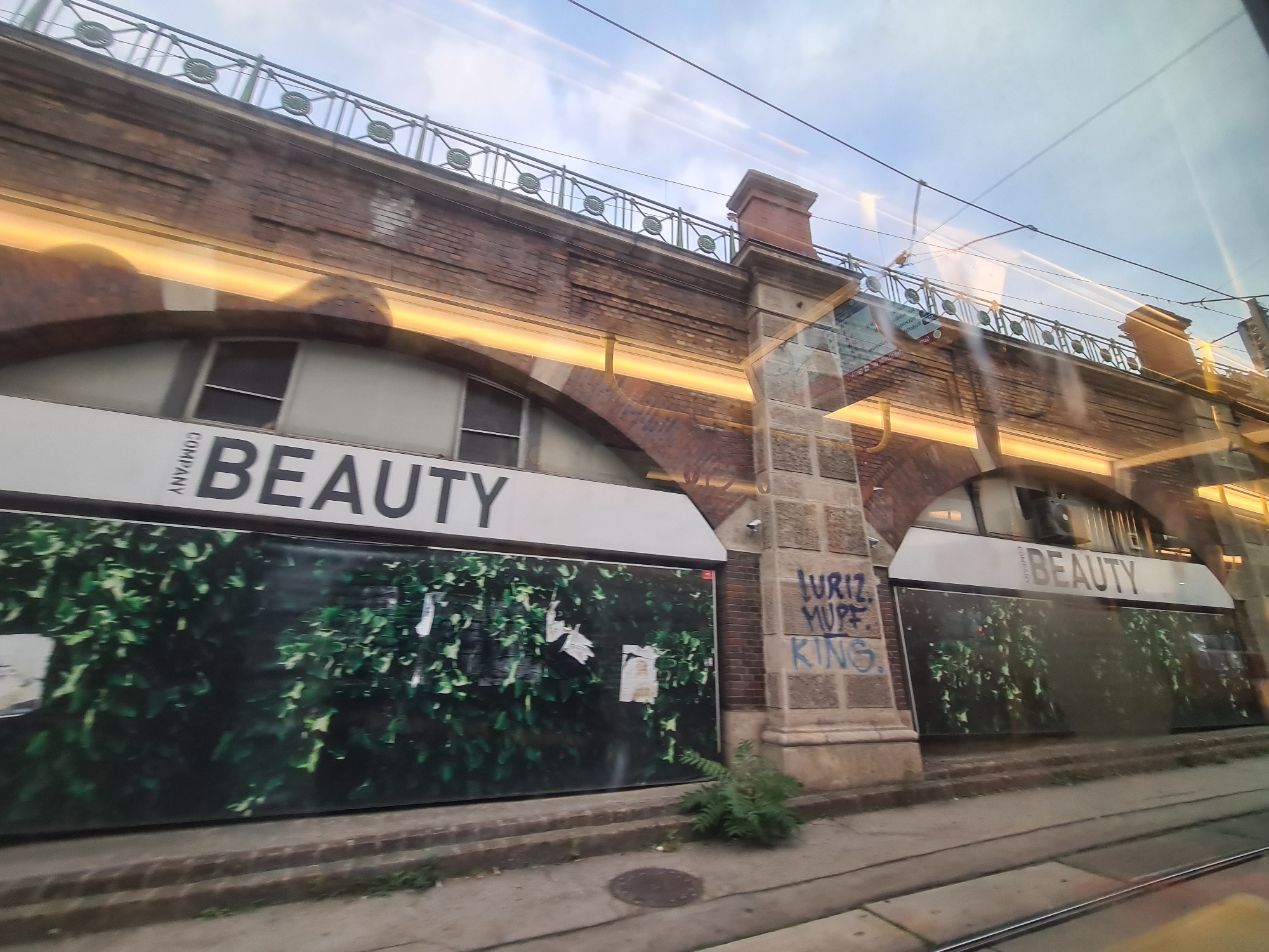 The image depicts a view from a tram window in Vienna, Austria. The tram is passing by an underpass structure, likely part of a railway or elevated road. The underpass is constructed of brick and stone, with arched openings. On the wall of the underpass, graffiti is visible, including the words "LURIZ", "MUPE", and "KING". Below the underpass, there are storefronts or boarded-up windows with large posters of green foliage. The word "BEAUTY" is prominently displayed in large, black lettering on white signage above these posters, with "COMPANY" written vertically to the left of "BEAUTY". The sky is visible in the background with some clouds and overhead power lines for the tram. The overall impression is of an urban environment, likely a transportation hub or a street with commercial activity.
