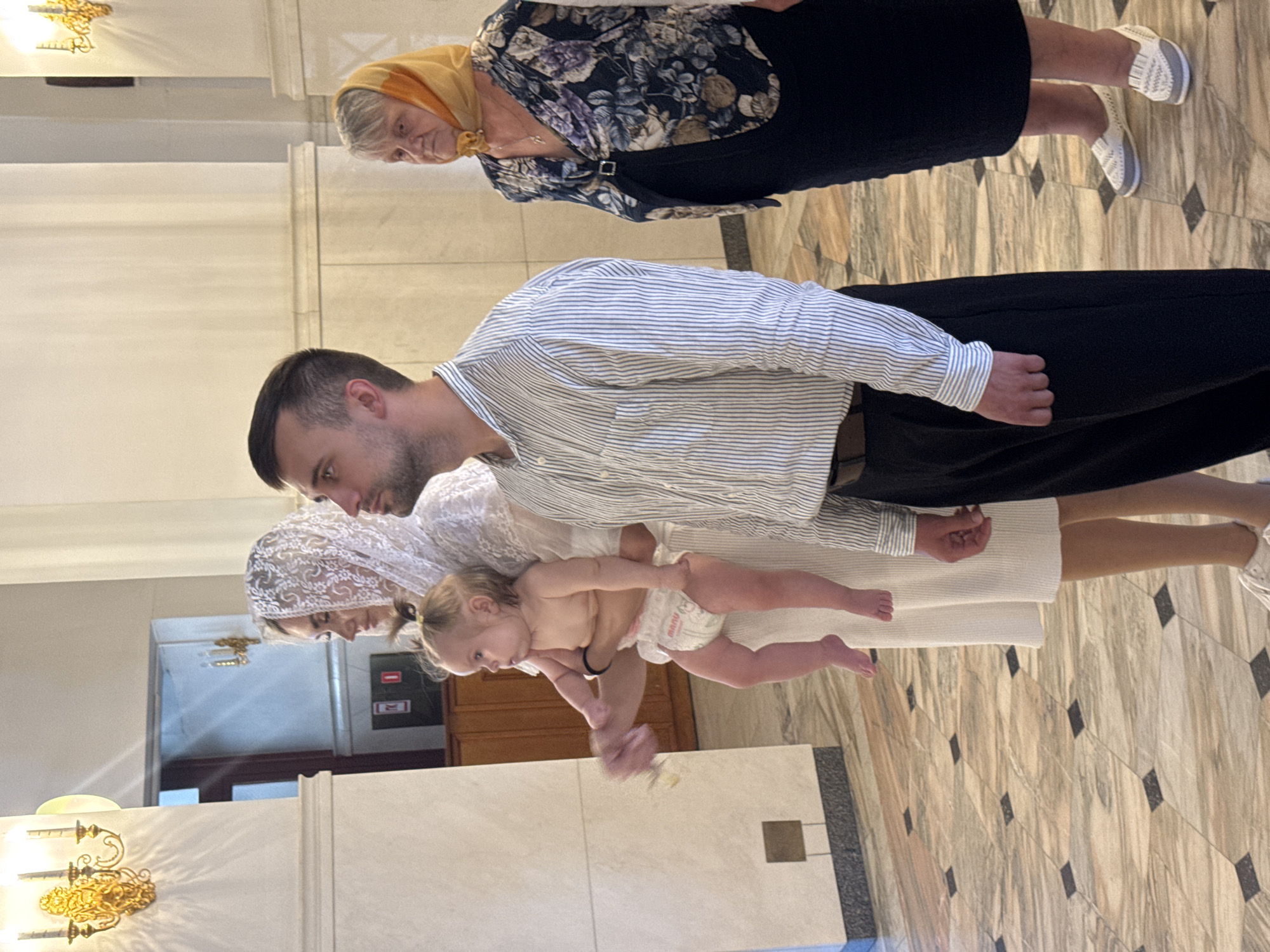 Four people are standing inside a building with a marble tiled floor and tall white columns. On the left, a woman wearing a white dress and a white lace veil is holding a baby. The baby is wearing a diaper and is facing to the left. Next to her, a man in a striped shirt and black pants is looking forward. To the right of the man, an older woman is wearing a floral patterned jacket and a yellow headscarf, looking to the left. The background shows wooden cabinets and sconces on the wall. The scene appears to be in a place of worship.