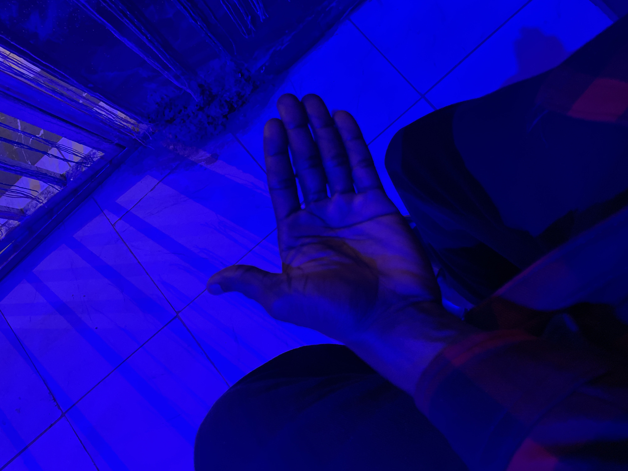 In Durumi, Nigeria, an individual is positioned with their left leg bent and their right hand extended forward, palm open. The person is wearing a checkered shirt and dark trousers, bathed in a vibrant blue light that casts strong shadows. The background consists of a tiled floor with a visible grate in the lower right quadrant of the image. The overall lighting creates a dramatic and somewhat abstract visual.