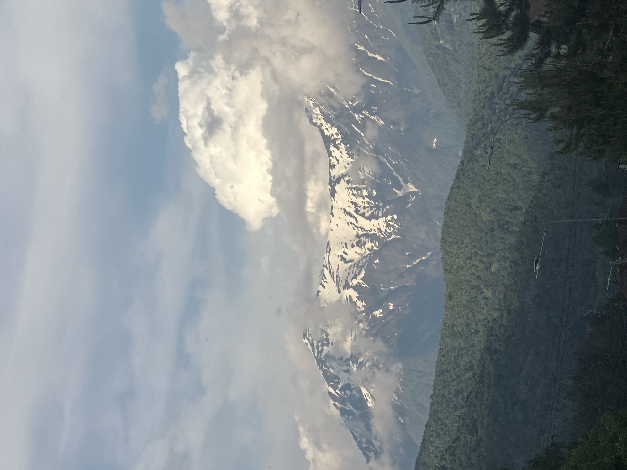 The image depicts a panoramic view of mountainous terrain, likely captured from the city of Krasnaya Polyana, Russia, looking towards the surrounding peaks. In the background, majestic mountains with significant snow cover are visible, indicating either high altitude or a specific season. The snow appears patchy, clinging to the higher elevations and crevices of the dark, rocky peaks. These snow-capped mountains are partially shrouded by voluminous white and grey cumulus clouds, with some clouds obscuring portions of the peaks and creating a dramatic atmospheric effect. A pale blue sky is visible above and between the clouds.
In the mid-ground, a dense expanse of lush green coniferous and deciduous forest covers rolling hills and lower mountain slopes, indicating a rich temperate or boreal ecosystem. The green foliage gradually transitions into the lighter, snow-dusted tones of the higher elevations.
The foreground is dominated by closer, darker green trees, suggesting a more immediate proximity to the viewer. Several man-made elements are present: a street light with an elongated, horizontal fixture is visible in the lower-middle portion of the image, extending from the left towards the center. Below it, multiple dark power lines or communication wires traverse horizontally across the frame. To the far right, the dark, spiky silhouettes of what appear to be pine trees are visible, along with a hint of a reddish-brown rooftop or structure in the very bottom right corner. The lighting suggests daytime, with diffused sunlight likely from behind or to the side of the viewer, illuminating the clouds and snow-capped peaks. No people are visible in the scene.
