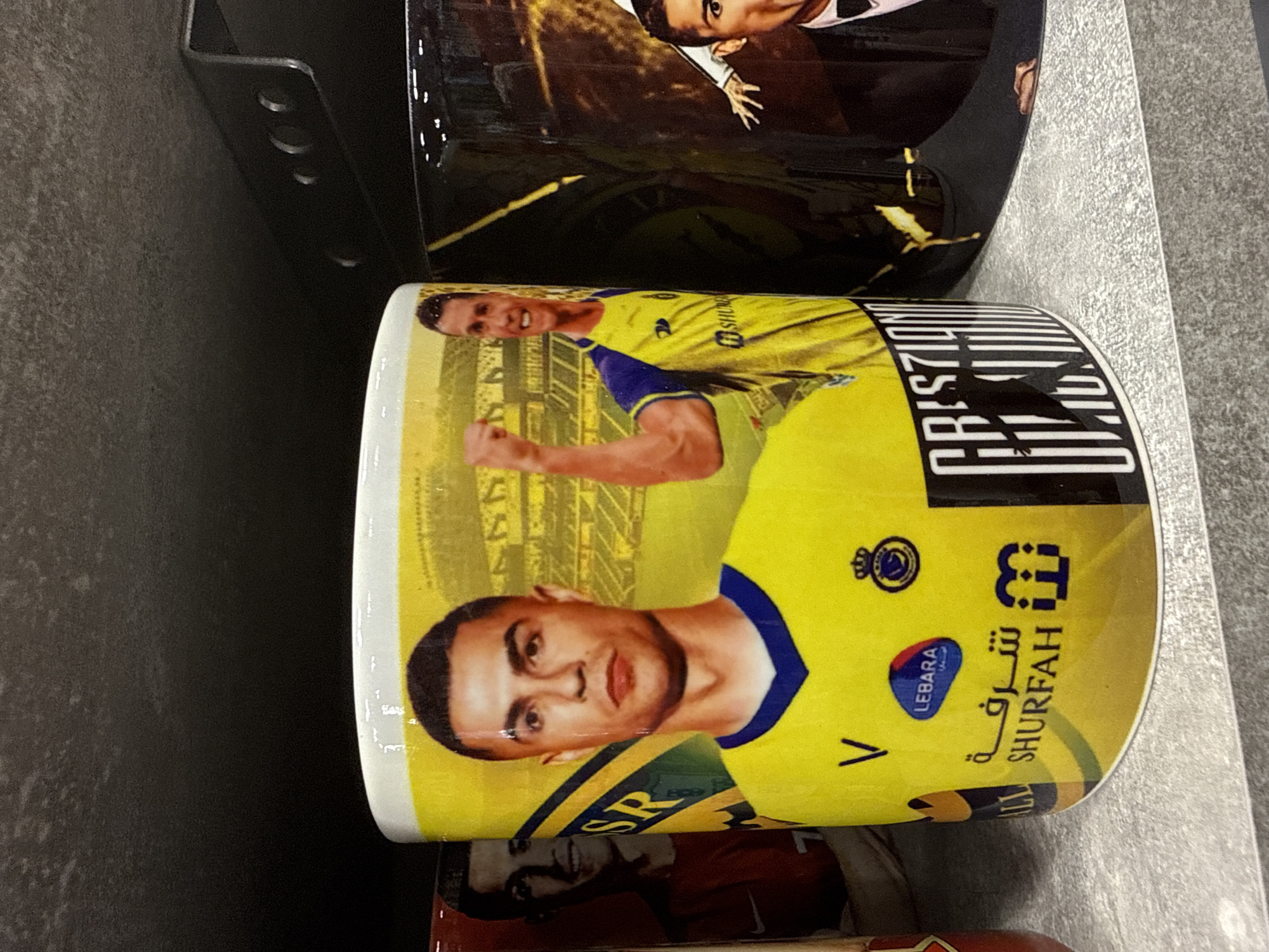 A close-up photograph displays three novelty mugs featuring images of Cristiano Ronaldo, arranged on a light gray, speckled shelf. The central mug, predominantly yellow, showcases a frontal portrait of Ronaldo in a yellow and blue Al Nassr football jersey, looking directly forward. To the right of this portrait, a smaller, full-body image of Ronaldo in the same jersey is depicted mid-celebration, with a fist raised and a stadium background. Below the main portrait, various logos are visible, including "LEBARA," Arabic script with "SHURFAH," and a crown emblem. On the far right side of this mug, vertical white text spelling "CRISTIANO RONALDO" is partially visible, overlaid with a black silhouette of Ronaldo. To the left, a portion of a reddish-brown mug with a faded image of Ronaldo in a red jersey and the number '7' is visible. To the right, a section of a dark-colored mug shows Ronaldo in a black tuxedo with a bow tie, accompanied by a partial image of a white hand. The background behind the mugs is a dark, textured surface, with a metallic bracket featuring three circular holes visible in the upper right. The scene is located in the city of Primorsko, Bulgaria.