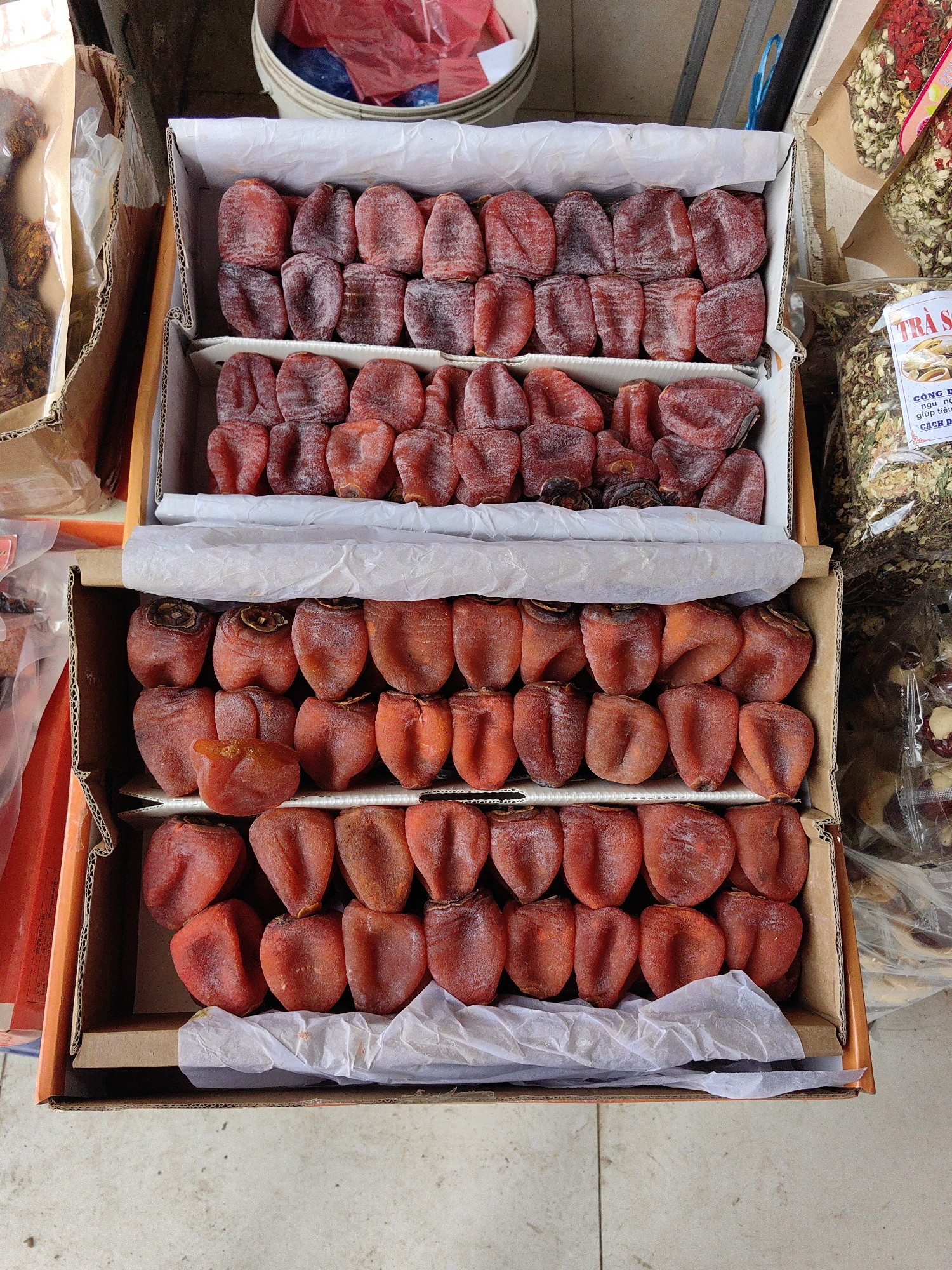 The image displays an overhead view of a market stall in Sa Pa, Vietnam, showcasing various dried food products. The central focus is a two-tiered arrangement of dried persimmons within stacked corrugated cardboard boxes. The top box contains two layers of shriveled, reddish-brown dried persimmons, some with a faint white powdery coating, arranged in neat rows. The bottom box similarly holds two layers of the same dried fruit. White crumpled tissue paper separates some of the persimmon layers within the boxes.
To the immediate left of the boxes, a brown paper bag contains a translucent plastic bag filled with dark, irregularly shaped dried items. Further left and above, a white plastic bucket holds red and blue plastic bags. To the right of the persimmon display, multiple clear plastic bags are visible, containing various dried herbs and floral components. One prominent bag features a white label with red and black Vietnamese text, partially visible as "TRÀ S..." (likely indicating "Sapa Tea" or similar). Other bags contain what appear to be dried jasmine flowers, red flower petals, and reddish-brown dried fruits or berries. The entire display rests on a light-colored, possibly tiled or concrete, ground surface. No individuals or specific actions are depicted.