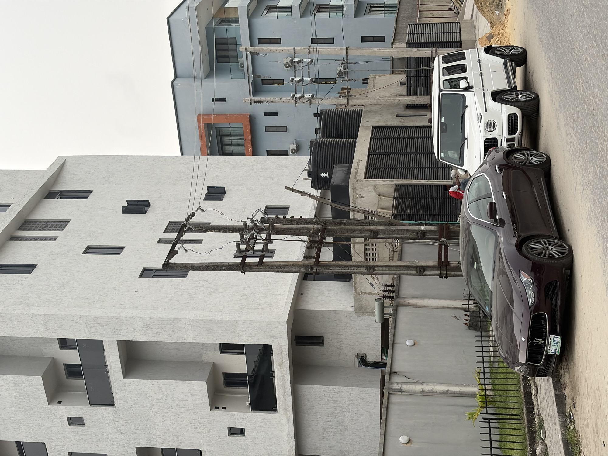 The image captures an urban street scene in what appears to be a residential or commercial area. On the left, a prominent modern building with a textured white facade stands tall, featuring numerous square and rectangular windows, some recessed. Behind it and to the right, another multi-story building with a light blue-grey exterior is visible, also exhibiting a modern architectural style with multiple windows and visible air conditioning units.
In the mid-ground, a dense network of utility poles and electrical wires stretches across the frame, indicating an active power infrastructure. Several black, cylindrical tanks (likely water storage) are situated on the ground level near these utilities, with one tank displaying a symbol resembling a stylized 'H'.
Parked on the right side of the street are two luxury vehicles. In the foreground, a dark-colored (possibly maroon or dark brown) Maserati sedan is visible, its front grille and headlights clearly in view. The license plate on this car reads "AGL 148GY" and "LAGOS," confirming the location as Lagos, Nigeria. Behind the Maserati, a white Mercedes-Benz G-Wagen SUV is parked. A person wearing a red shirt is partially visible behind the Maserati, near the G-Wagen.
The day appears to be overcast or cloudy, as indicated by the diffused lighting and lack of sharp shadows. A paved road occupies the right side of the image, while a short metal fence and some sparse green plants are seen along the roadside. The overall scene depicts a blend of modern architecture and infrastructure common in urban Nigerian settings.