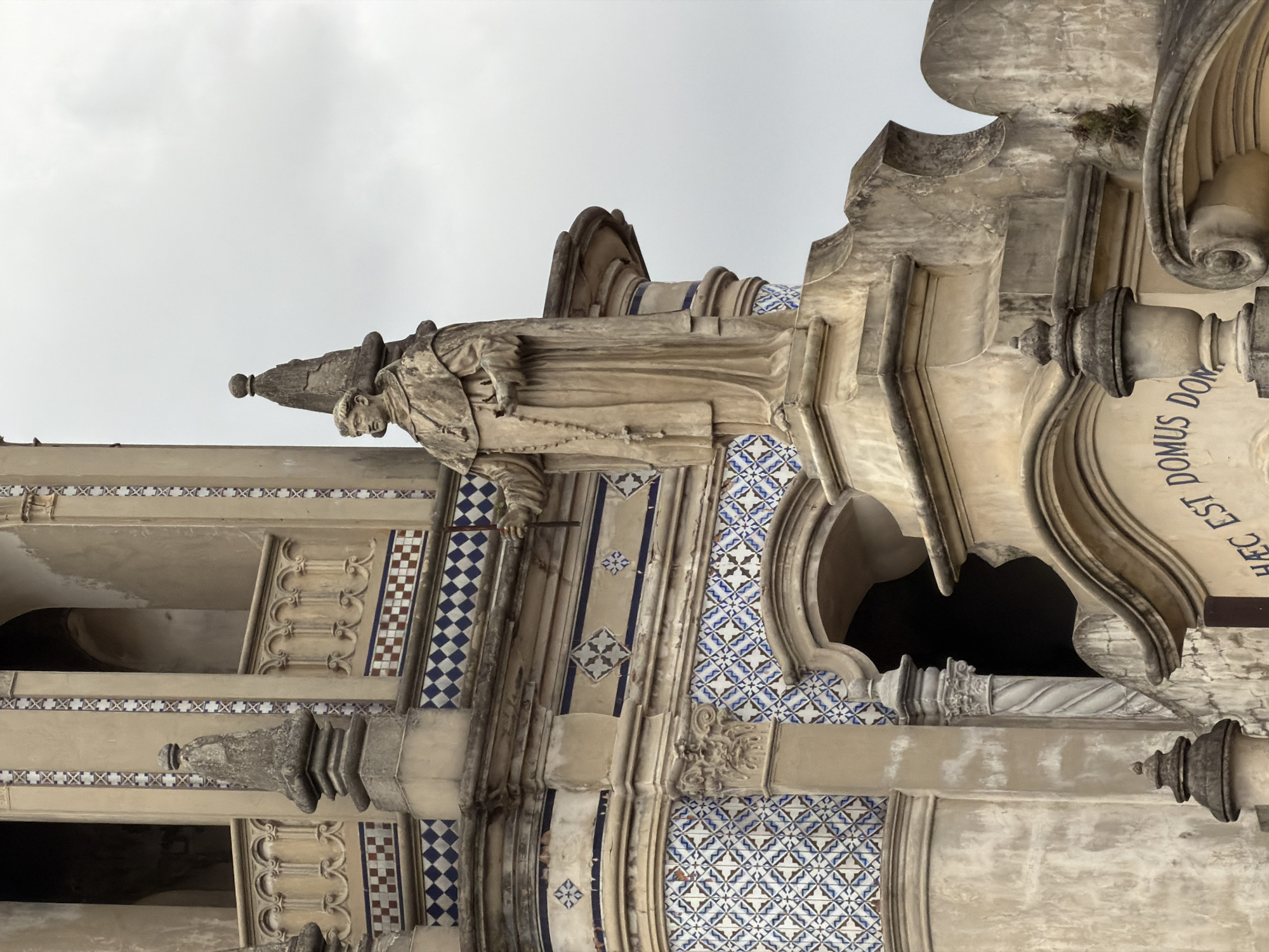 The image shows a section of a building facade in Buenos Aires, Argentina. The architectural style is ornate, featuring detailed stonework, a statue of a monk holding a rosary, and decorative blue and white tiles. The building's facade also includes a stone structure that contains the words "HEC EST DOMUS DOM." The sky is overcast, suggesting a cloudy day.
