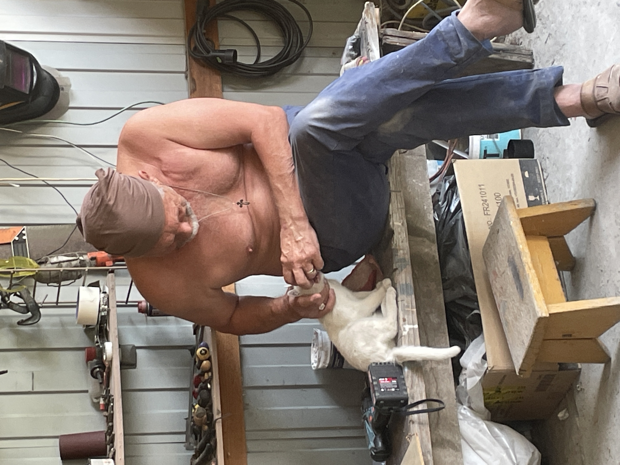 A shirtless man, wearing a brown bandana on his head and blue jeans, is sitting on a wooden bench in what appears to be a workshop or garage. He is interacting with a white cat, gently holding its head with his hands. Tools and equipment are visible on shelves behind him, including a welding mask hanging on the wall. Various boxes and a small wooden stool are on the floor in front of the bench. The environment suggests a practical, working space.