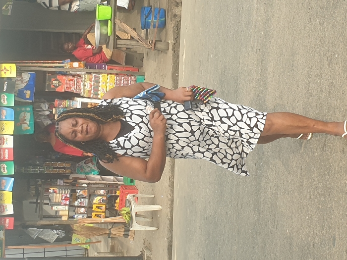 This image captures a daytime street scene in Umuode, Nigeria, featuring a woman walking on an asphalt road in the foreground, with a roadside shop or stall in the background.
The primary subject is a woman of medium complexion, walking towards the viewer. She has dark hair styled in braids with visible lighter streaks and wears a sleeveless dress with a distinctive black and white abstract pattern over a black undershirt. Her footwear consists of white flip-flops. Her expression appears solemn or serious. In her left hand, she holds a crumpled light blue cloth. In her right hand, she carries a small, multicolored beaded clutch purse alongside a dark rectangular object, possibly a mobile phone.
The background consists of a storefront. Above the entrance, various product advertisements for brands such as Milo, Nescafe, Quality, Nido, Johnny, Indomie, and Maggi are displayed. The shop entrance reveals assorted merchandise, including shelves stocked with what appear to be motor oils or lubricants in yellow bottles, bundles of brooms, and stacks of packaged beverages in crates. A small, white plastic table to the left holds a bunch of green bananas. Inside the shop, a person in a red shirt is partially visible. To the right, a man in a red shirt is seated on a low wooden bench next to a larger wooden table that holds a large metal bowl and a green plastic bucket. A third individual, wearing a striped shirt, is partially visible standing on the far right. The overall environment suggests a commercial area along a main thoroughfare.