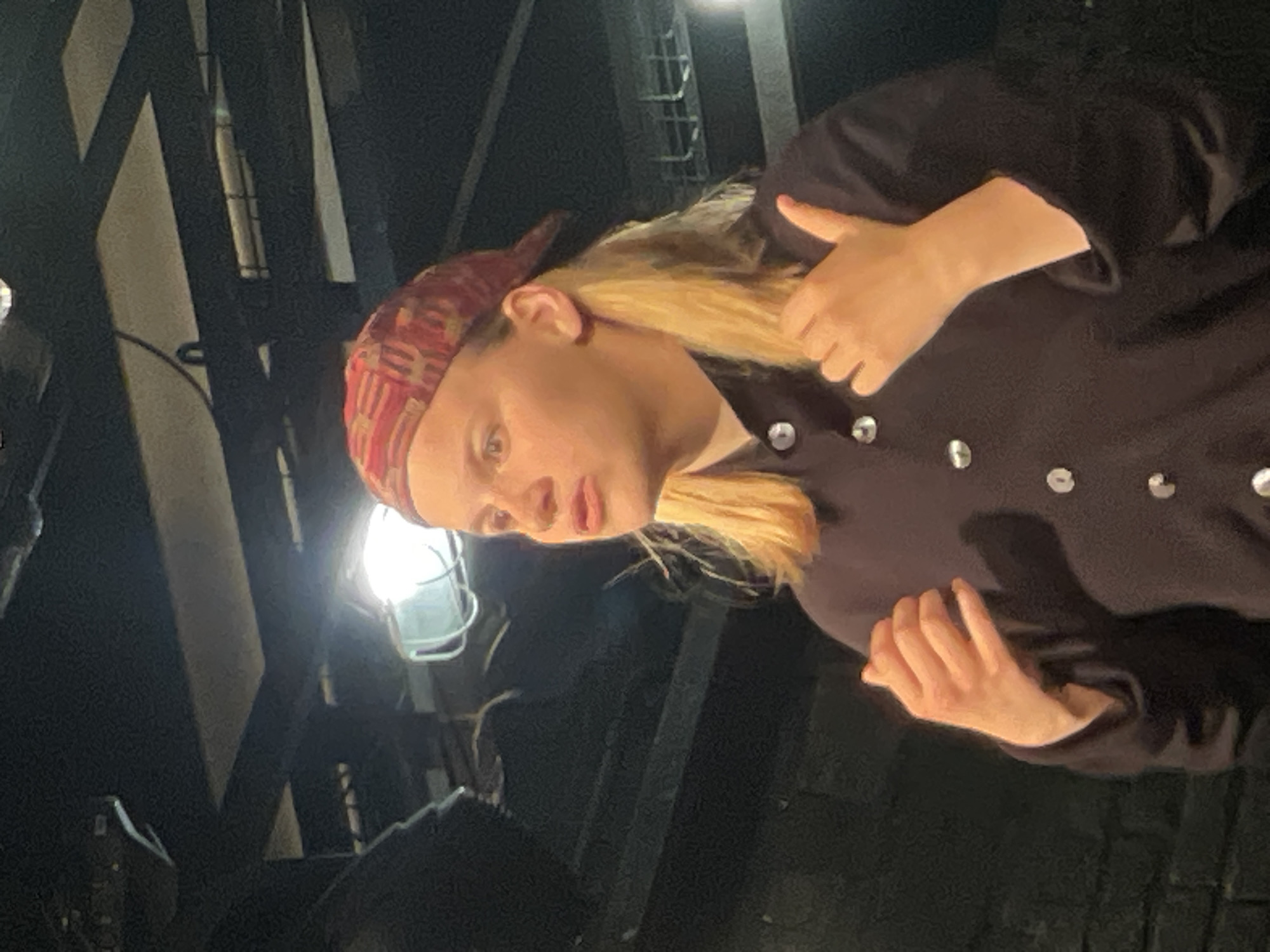 A medium-shot portrait captures a person, likely a woman with blonde hair, looking slightly off-camera with an engaged or contemplative expression. She is wearing a colorful red, black, and white geometrically patterned headscarf and a dark, long-sleeved top adorned with multiple circular silver buttons down the front. Her mouth is slightly open, suggesting she might be speaking or reacting, and her left hand is visible near her waist, fingers gently curled.
The background is dark and appears to be an indoor setting, likely an industrial space, a theater backstage, or a workshop. This type of utilitarian environment could be found in Moscow, Russia. Bright, exposed utility lights with protective cages are visible, casting strong, focused illumination on the subject's face and upper body, while much of the background remains in shadow. Behind her, dark, intricate metal structures, resembling scaffolding or pipes, are dimly visible. The overall atmosphere is dark, with artificial light highlighting the subject. There is no visible text in the image. The image is captured in a rotated orientation (90 degrees clockwise from portrait).