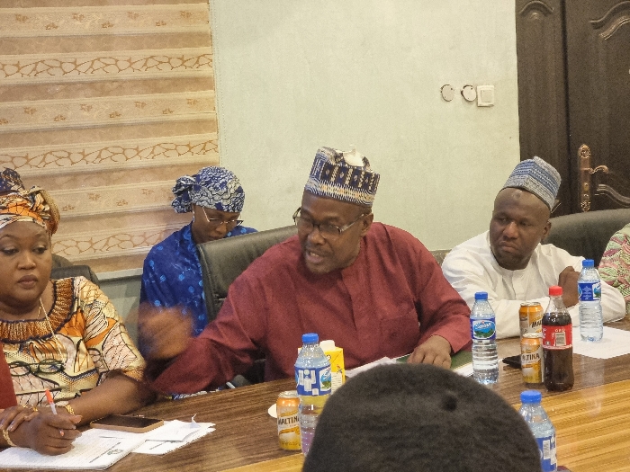 The image shows four people sitting around a table in an indoor setting. The table is covered in a wood veneer, with a variety of food and drink items scattered across its surface. Two of the people are wearing traditional Nigerian clothing and are sitting in chairs that are visible in the image. Another person is wearing a maroon long-sleeve shirt, and a fourth person is wearing a blue dress. They appear to be engaged in conversation. The setting is likely an office or meeting room in Maiduguri, Nigeria. The coordinates provided (latitude: 11.832, longitude: 13.143) correspond to a location within Maiduguri.