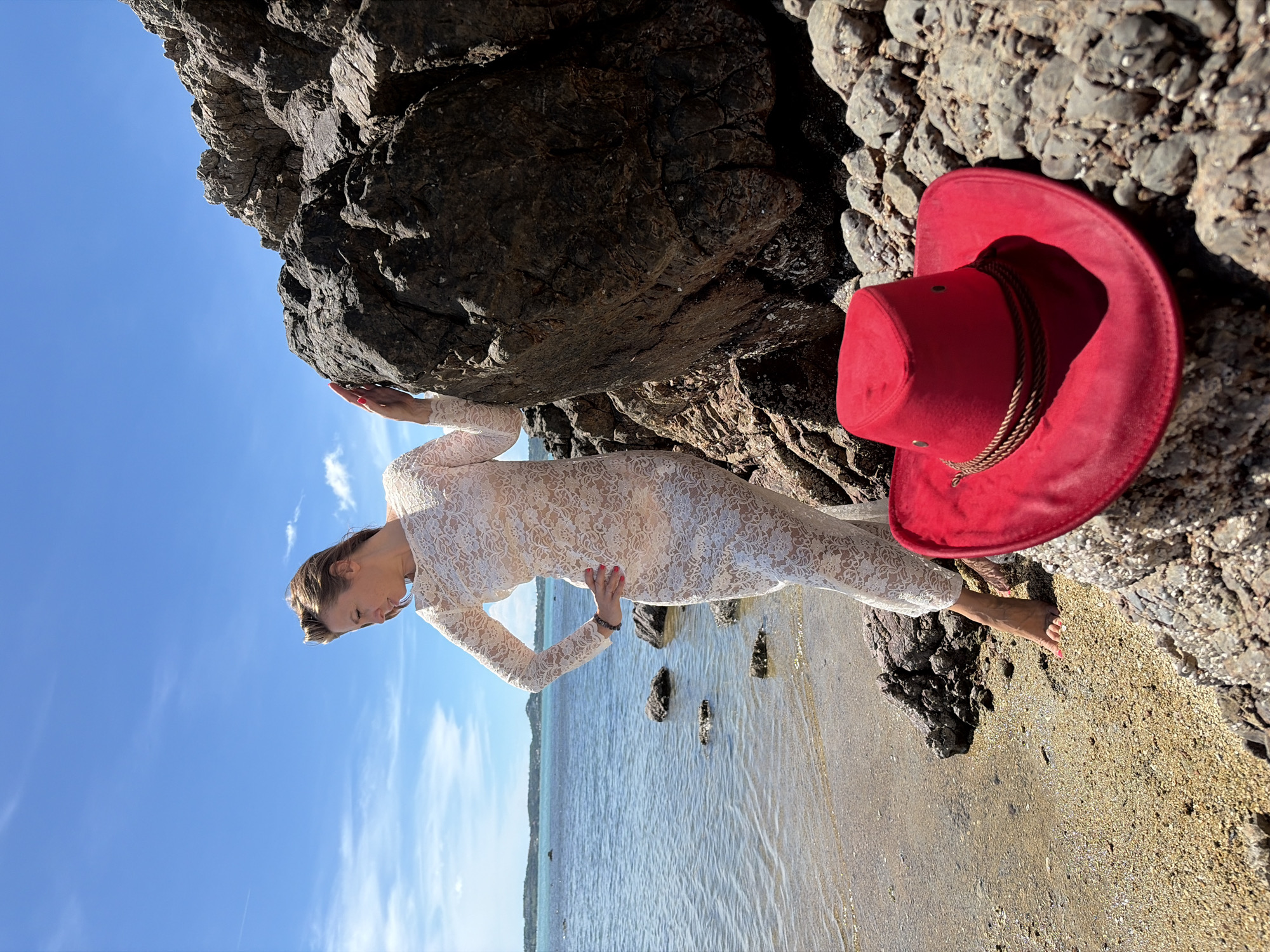 In Baan Bo Phut, Thailand, a woman in a white lace dress stands on a rocky beach with the ocean to her left and a large rock formation to her right. She is looking down and to her left, with her left hand on her hip and her right hand resting on the large rock. In the foreground, a red cowboy hat rests on the rocks. The sky is blue with scattered clouds, and the water is calm.